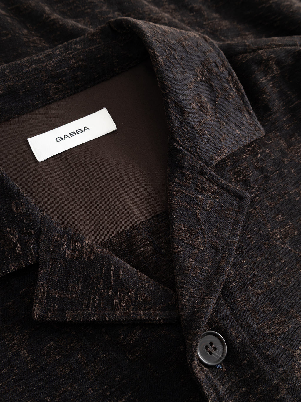 Close-up of a dark brown textured GABsimone Pockt Monte LS Overshirt - Brown Slate by Gabba, featuring a visible collar, black button, and a white label with the brand name inside the collar. This relaxed fit overshirt is a contemporary statement piece for any wardrobe.