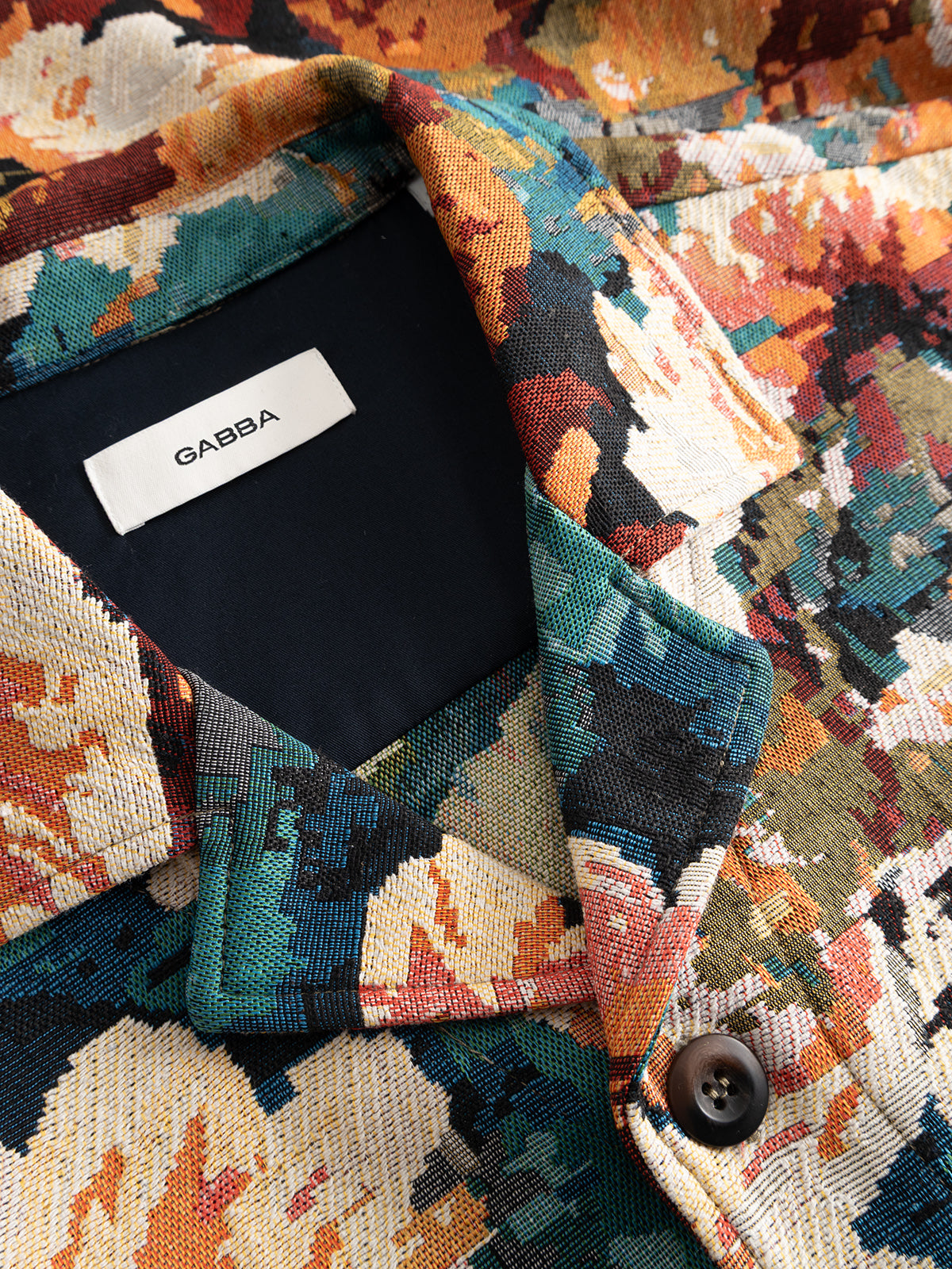 A close-up of the GABsimone Reso Fiore LS Overshirt by Gabba showcases its colorful, abstract multi-patterned fabric with vibrant splashes of orange, teal, cream, and black. A visible black label reading “GABBA” is sewn inside the collar, while the relaxed fit and bold design evoke the lively feel of a floral shirt.