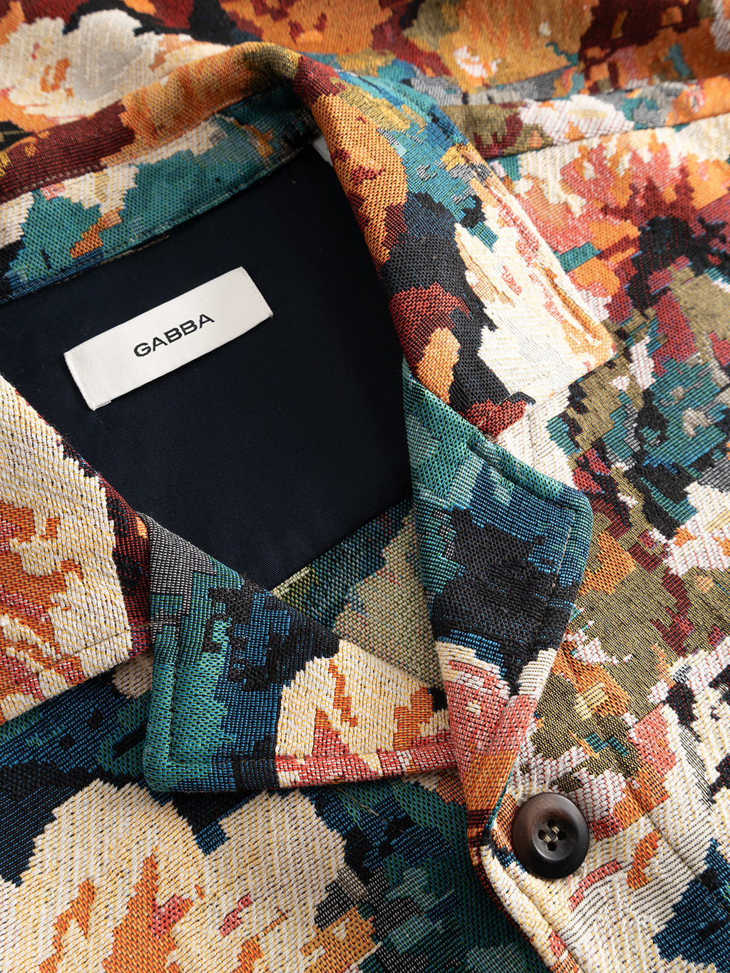 A close-up of the GABsimone Reso Fiore LS Overshirt by Gabba showcases its colorful, abstract multi-patterned fabric with vibrant splashes of orange, teal, cream, and black. A visible black label reading “GABBA” is sewn inside the collar, while the relaxed fit and bold design evoke the lively feel of a floral shirt.