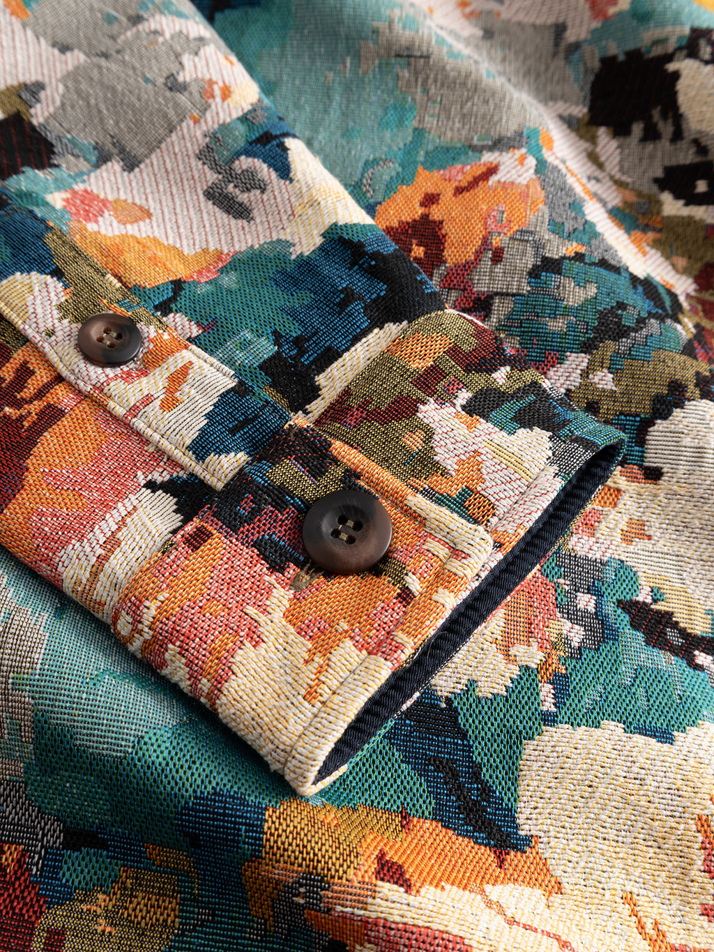 Close-up of the GABsimone Reso Fiore LS Overshirt by Gabba, showcasing a relaxed fit sleeve with a colorful abstract floral pattern and two dark brown buttons at the cuff. The viscose-blend jacquard fabric displays a textured weave in shades of teal, orange, yellow, black, and cream.