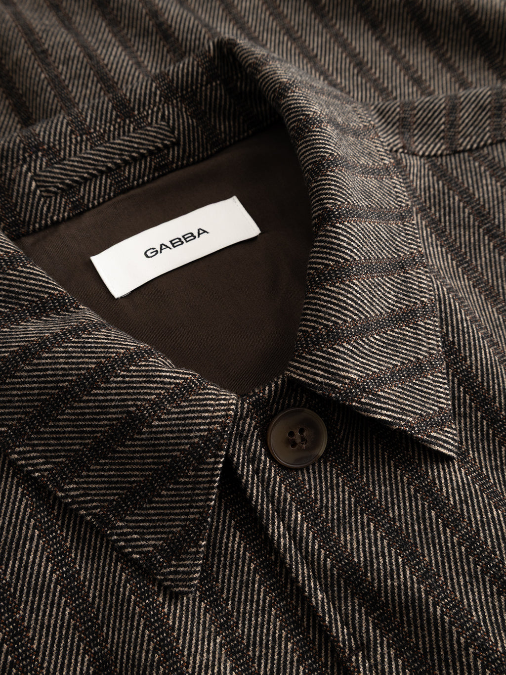 Close-up of the GABlibre AX Zapies LS Overshirt in Brown Slate, featuring brown and beige stripes, a collared neckline with button closure, chest pocket, and a white label inside the collar displaying the Gabba brand name. Designed in a regular fit, this lightweight overshirt offers both style and versatility for effortless layering.