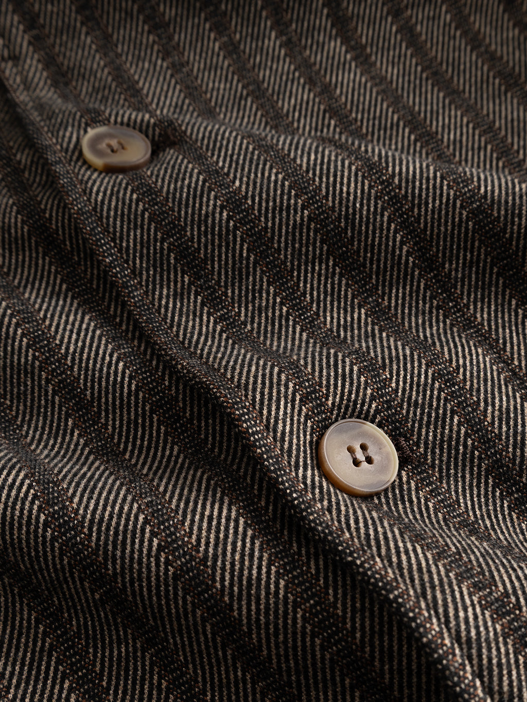Close-up of the GABlibre AX Zapies LS Overshirt - Brown Slate by Gabba, showcasing its fabric with dark and light vertical stripes and two brown buttons sewn onto the material. The clearly visible texture and stitching details highlight its quality, making it ideal for lightweight layering.