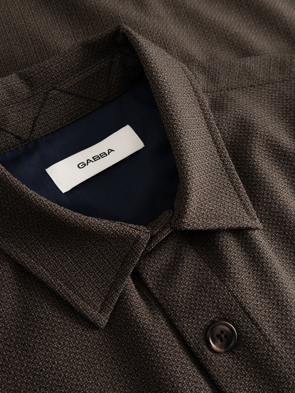 Close-up of the GABjens Over Rubin LS Overshirt in Brown Slate by Gabba, showcasing its textured stretch dobby fabric, a button closure, and a white label inside the collar reading Gabba. This versatile overshirt blends comfort and style with its subtle texture and premium finish.