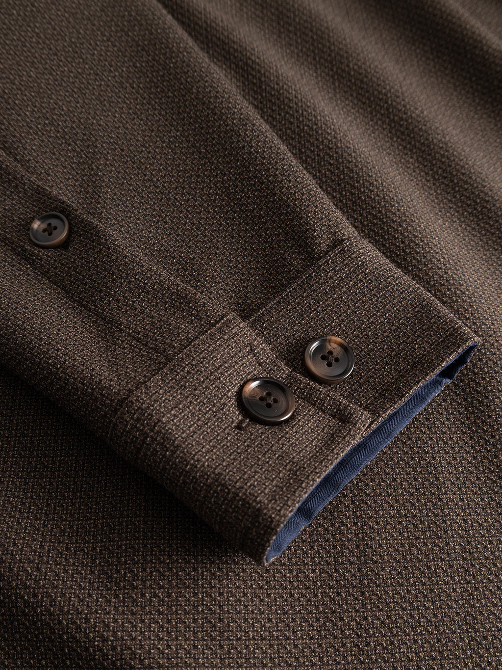 Close-up of the Gabba GABjens Over Rubin LS Overshirt in Brown Slate showcases the textured cuff constructed from stretch dobby fabric, featuring two dark buttons, visible stitching details, and a glimpse of dark blue inner lining—all lying flat on a surface.