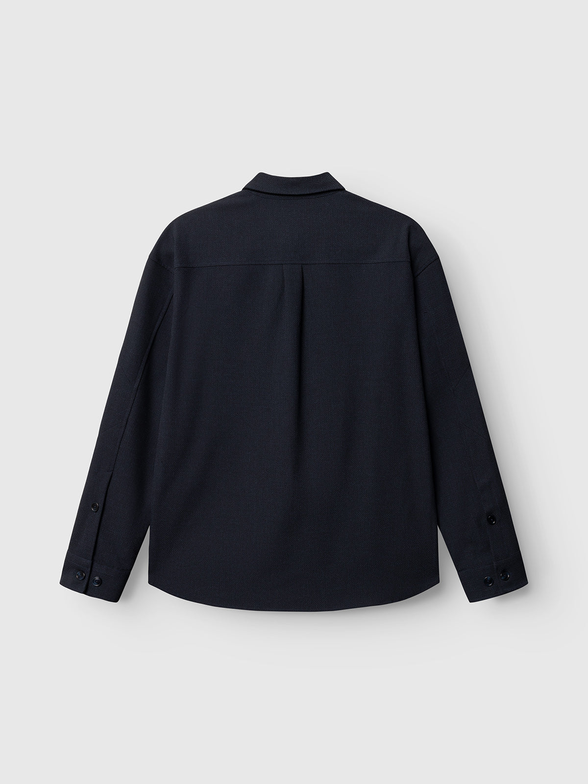 The GABjens Over Rubin LS Overshirt by Gabba in blue black is displayed from the back against a plain light grey background. This oversized, long-sleeve button-up shirt features a collar, buttoned cuffs, and a straight hem.