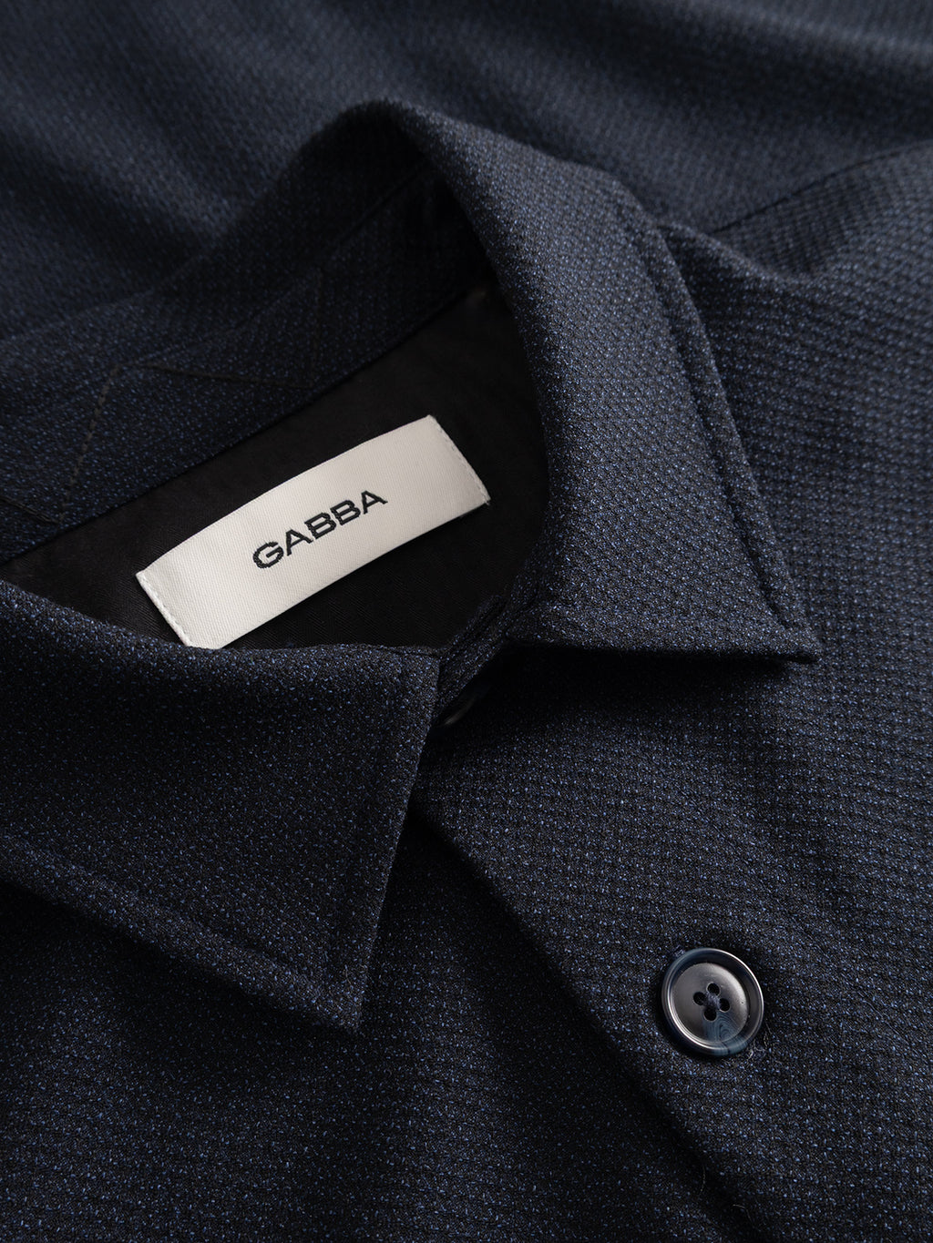 Close-up of the GABjens Over Rubin LS Overshirt by Gabba in blue black, featuring a pointed collar and crafted from stretch dobby fabric. The label inside the collar displays the Gabba brand, while a single matching button in the foreground highlights its relaxed overshirt style.
