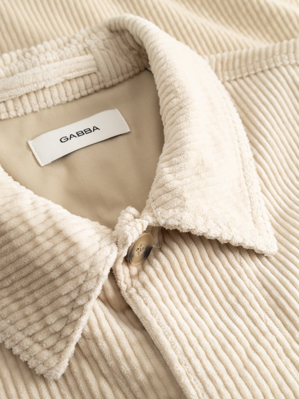 Close-up of the GABbeta Ibre LS Overshirt in Birch by Gabba—a cream-colored stretch corduroy shirt featuring a prominent collar, top button, and a white “Gabba” label inside the neckline—an elevated layering essential.
