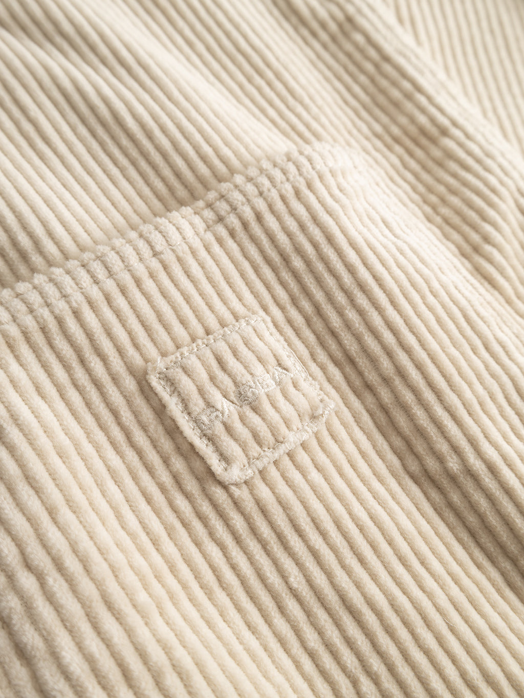 Close-up of the GABbeta Ibre LS Overshirt in Birch by Gabba, featuring cream-colored stretch corduroy fabric with vertical ridges and a stitched square label. The soft, plush texture makes it an ideal layering essential.