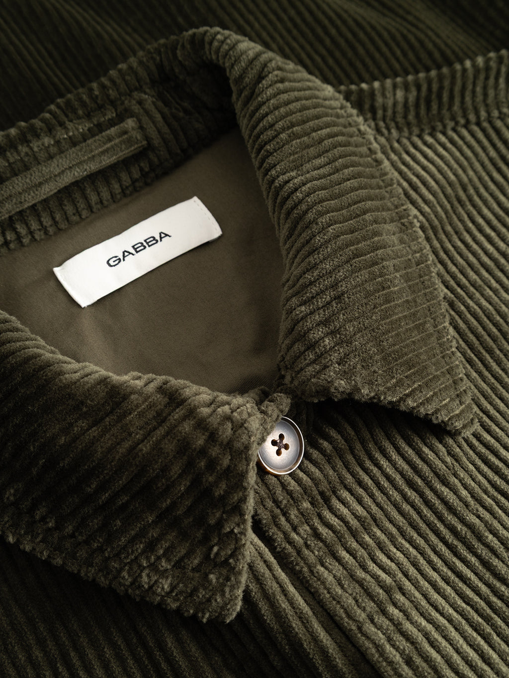 Close-up of the GABbeta Ibre LS Overshirt by Gabba in Forest Night—a stretch corduroy shirt featuring a visible collar, single button closure, and an inner white label with the Gabba logo—making it an ideal layering essential for any wardrobe.