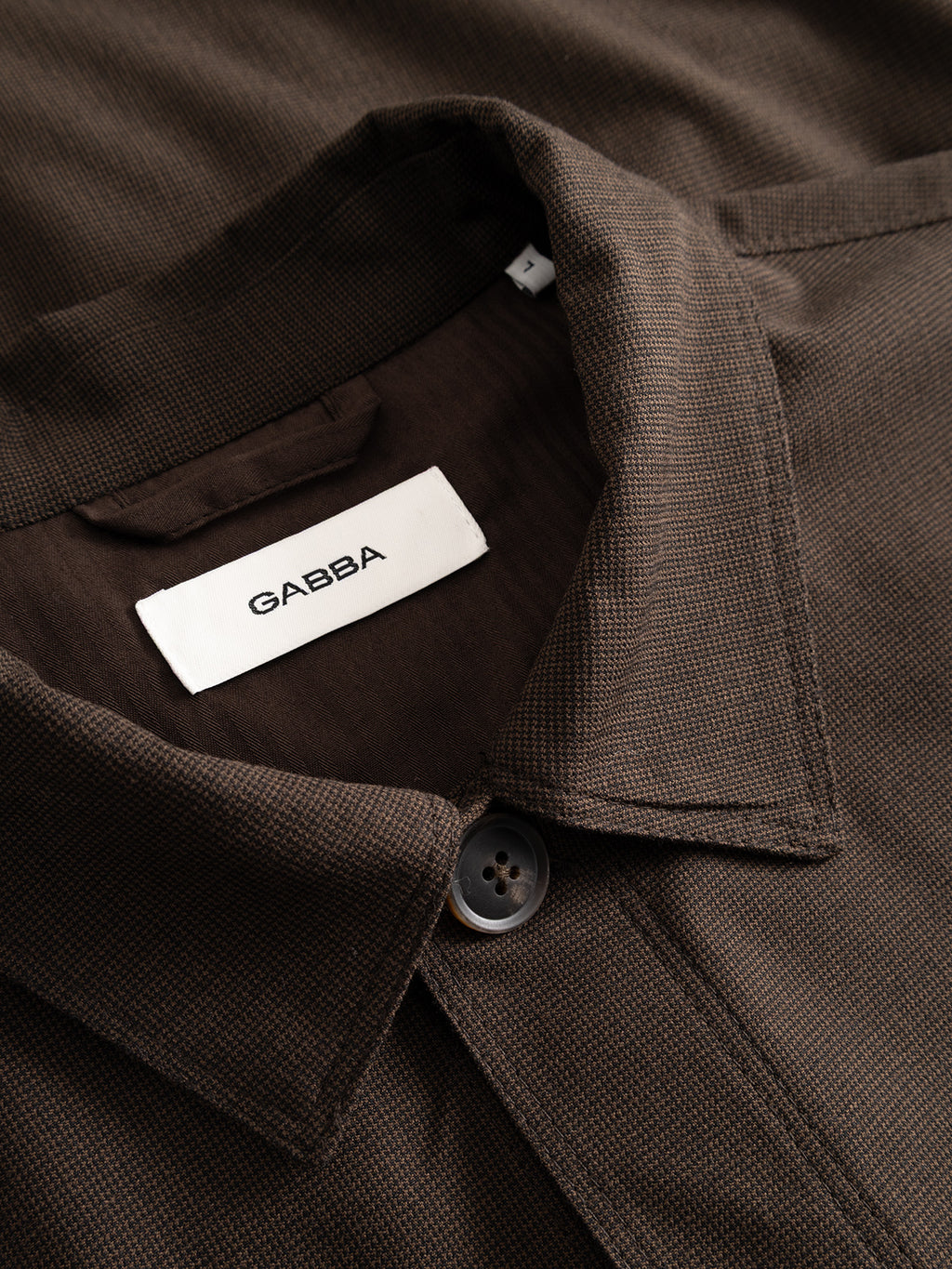 Close-up of the GABtogger Base Deme LS Overshirt in Brown Slate from Gabba, featuring a lightweight overshirt design with a brown collar, a visible black button, and an inner white label displaying the Gabba brand. The micro-pattern woven fabric offers a subtle textured finish, highlighting its regular fit and modern style.