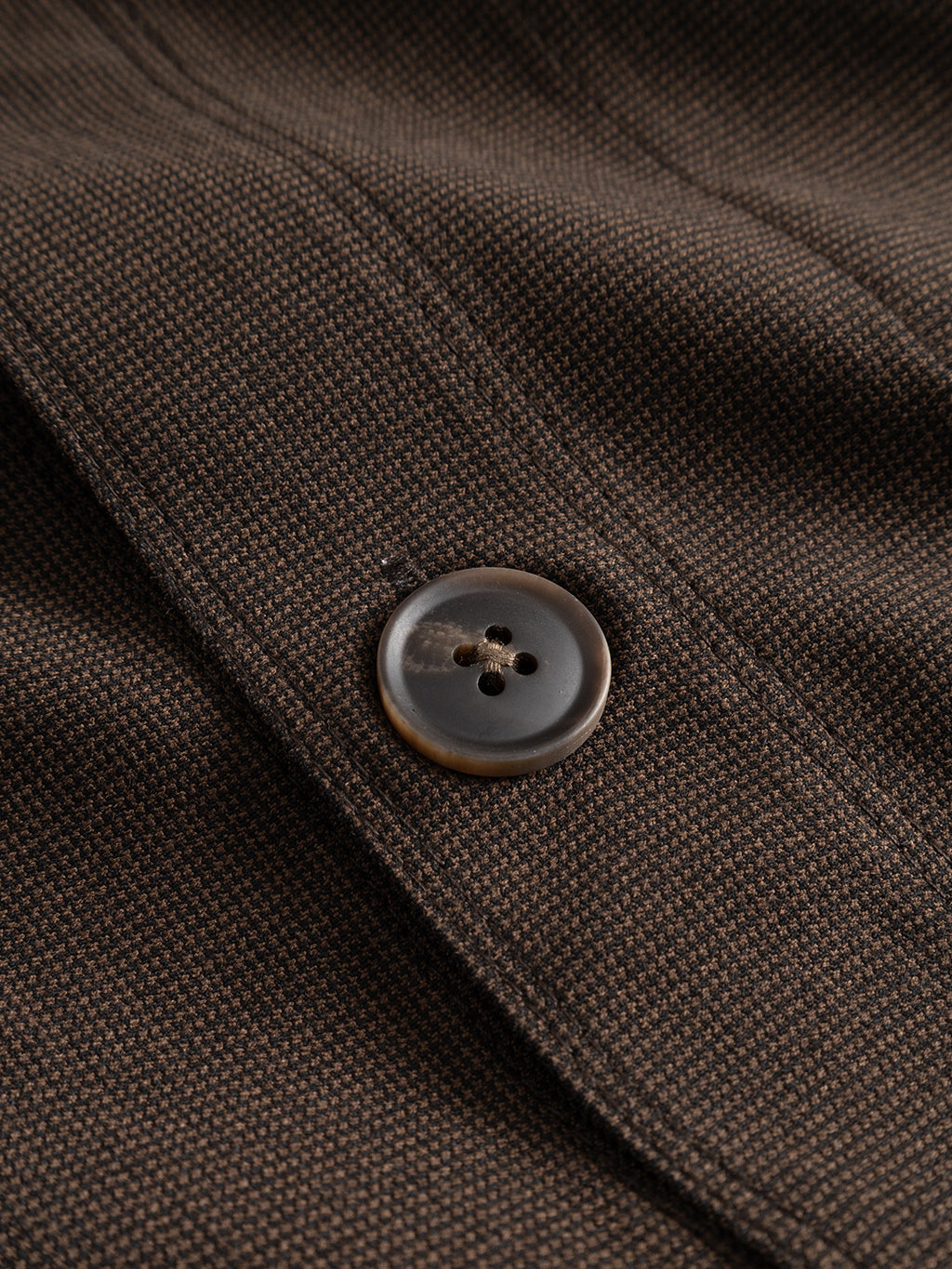 Close-up of a brown button sewn onto the textured, dark brown micro-pattern woven fabric of the Gabba GABtogger Base Deme LS Overshirt in Brown Slate. The visible stitching and detailed fabric pattern showcase the regular fit and meticulous craftsmanship.