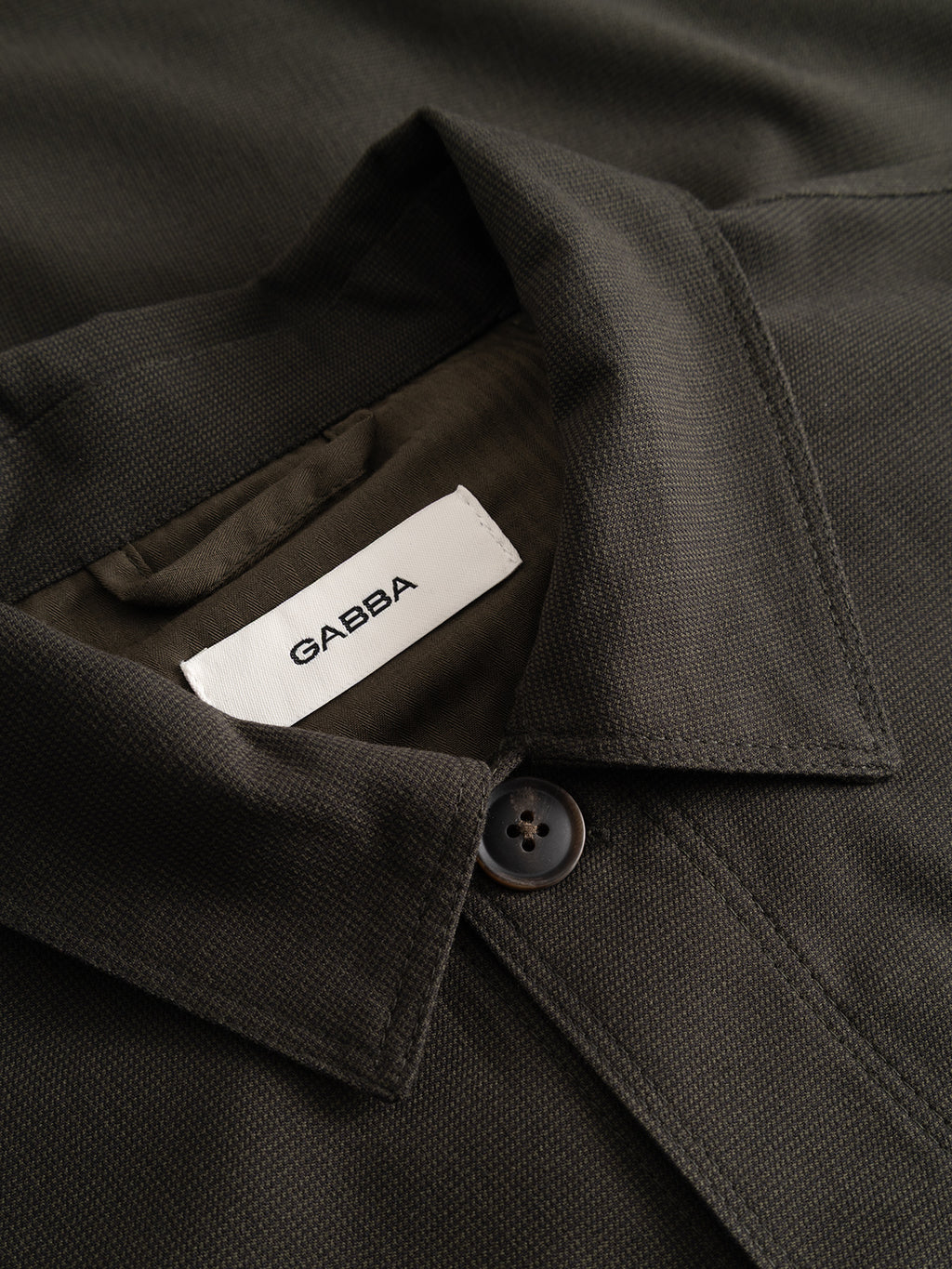 Close-up of the GABtogger Base Deme LS Overshirt in Forest Night, crafted from dark micro-pattern woven fabric and featuring a collar, a button, and a white label with the Gabba brand name sewn inside.
