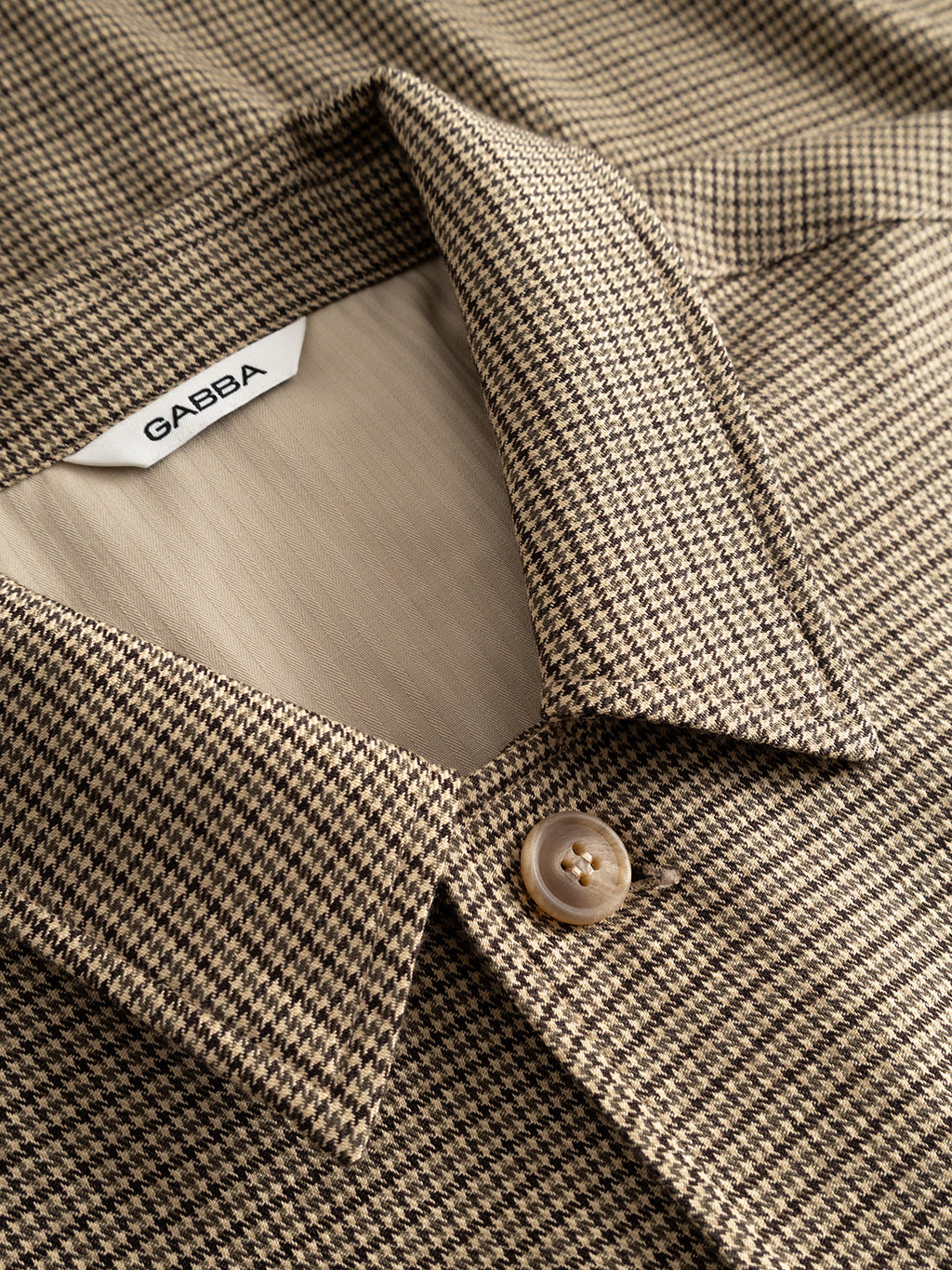 Close-up of the GABfelix Brit LS Shirt - Dark Sand by Gabba, a relaxed fit shirt made from lightweight woven fabric with a subtle beige and brown houndstooth pattern, finished with a beige button and an inner collar displaying the Gabba brand label.