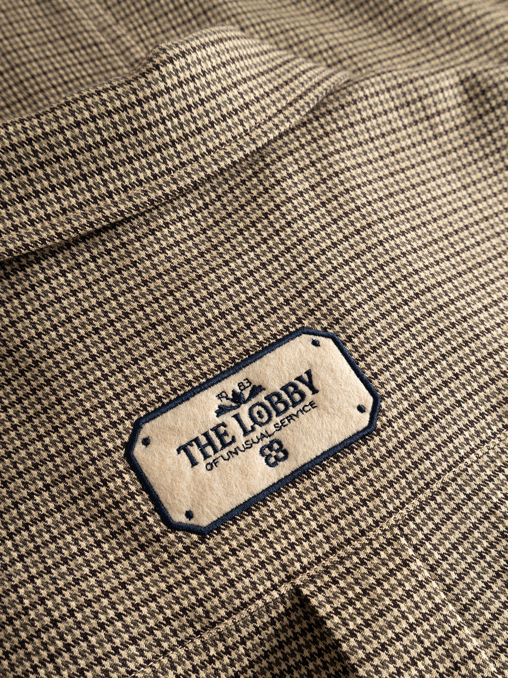 A close-up of the GABfelix Brit LS Shirt - Dark Sand by Gabba, featuring a houndstooth-patterned, lightweight woven fabric with a beige label that reads “THE LOBBY OF UNUSUAL SERVICE” in dark blue text, accented by small decorative elements and an embroidered border.