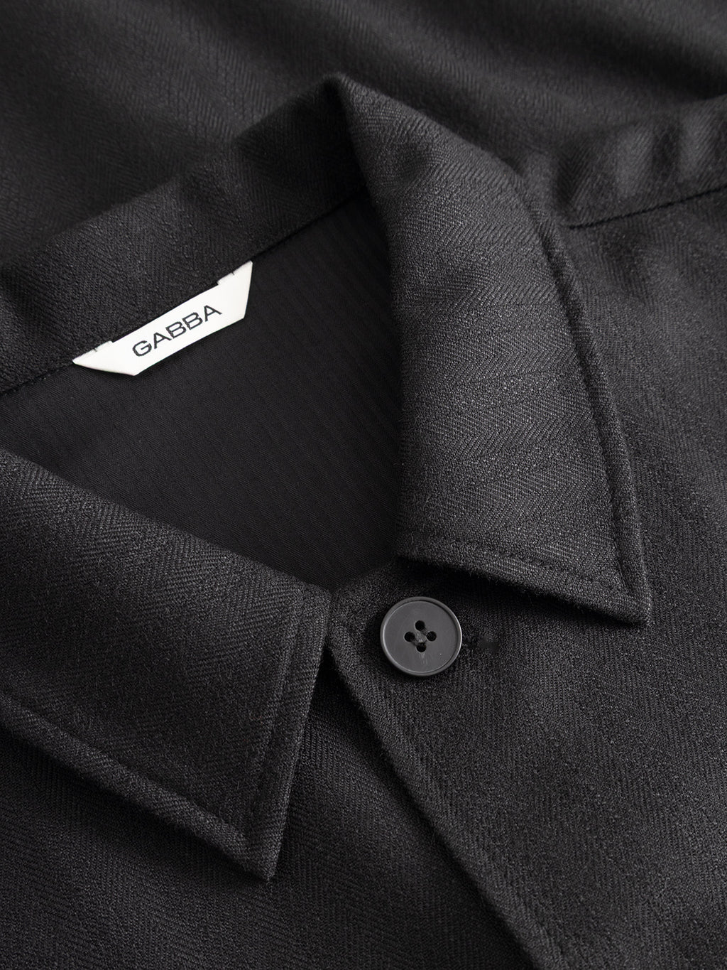 Close-up of the GABfelix Fuyu LS Shirt - Meteorite by Gabba, a black collared shirt with a single visible button and a white GABBA label inside the collar. Crafted from a viscose-blend fabric with a subtle textured pattern, this shirt offers a relaxed fit and comfortable everyday wear.