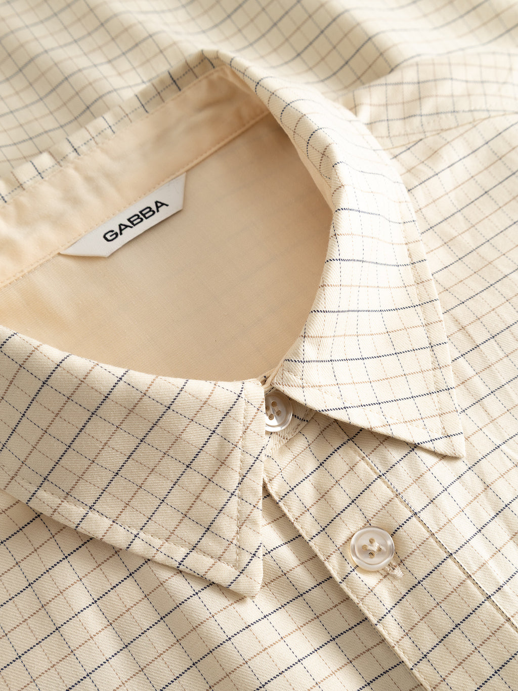 Close-up of the Gabba GABjens Over Indie LS Shirt in Lt. Sand, featuring a beige grid pattern and white buttons. Crafted from a soft cotton-blend fabric, this yarn-dyed checked shirt is neatly laid out with slight wrinkles, and the inner collar label displays the Gabba branding.
