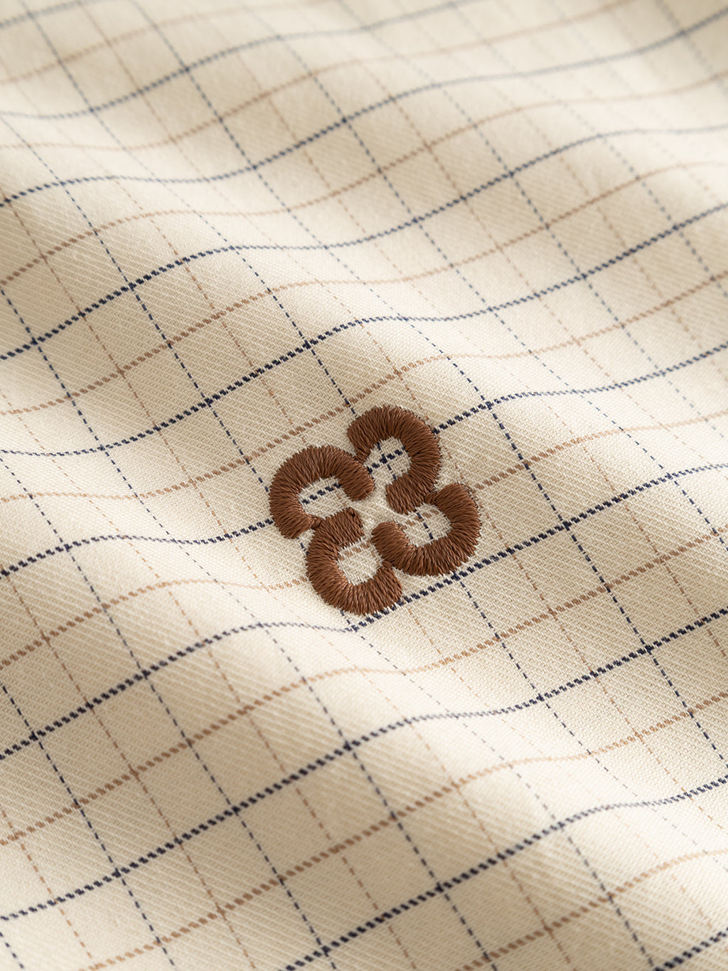 A close-up of the Gabba GABjens Over Indie LS Shirt in Lt. Sand, a yarn-dyed checked shirt made from a cream cotton-blend fabric with a blue and tan grid pattern, featuring a brown embroidered geometric design in the center.