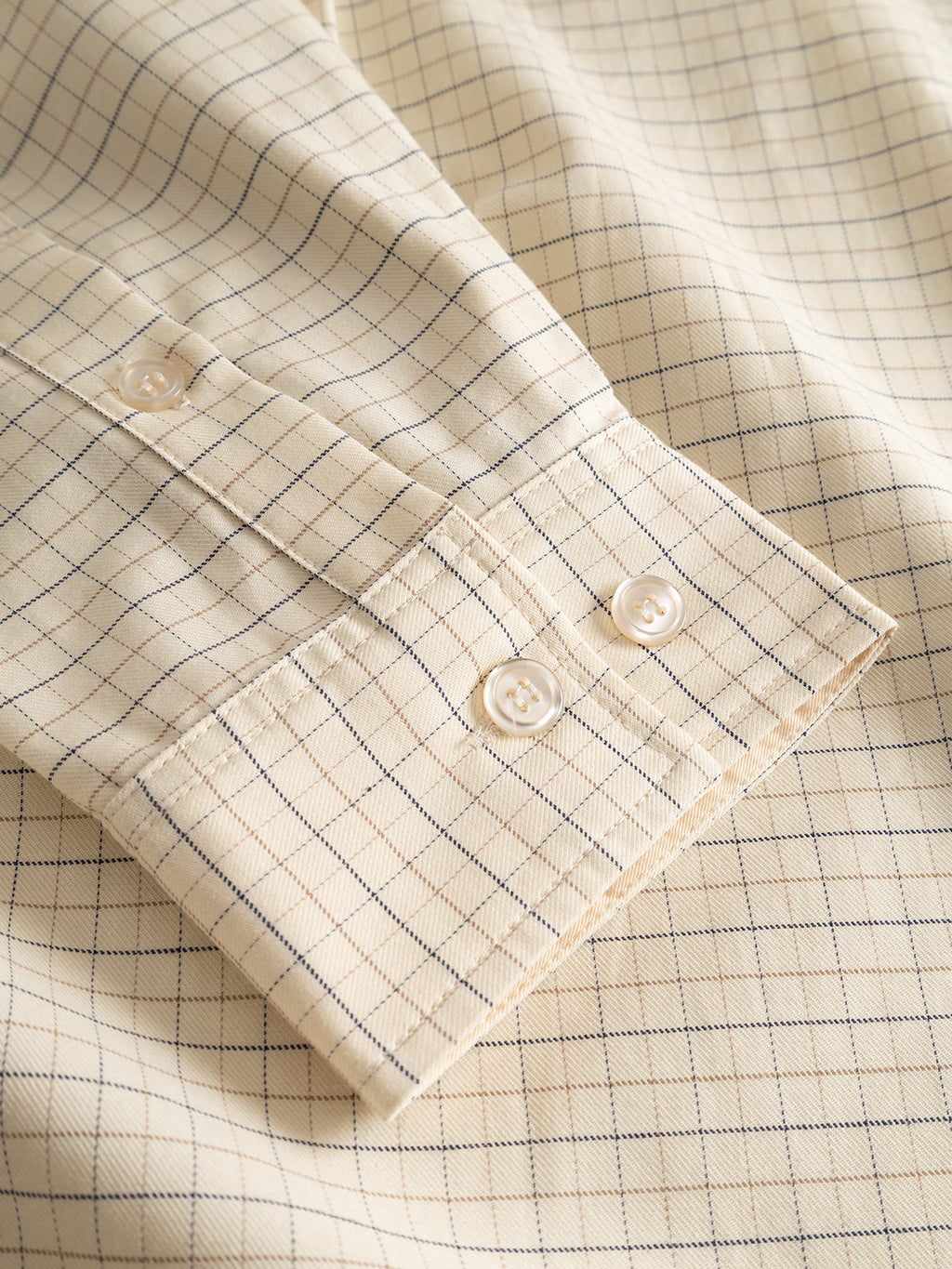 A close-up of the GABjens Over Indie LS Shirt by Gabba in light sand, featuring a cream-colored cotton-blend fabric with a blue and beige checkered, yarn-dyed pattern. The buttoned cuff has two white buttons, highlighting the shirt’s soft, slightly textured finish.