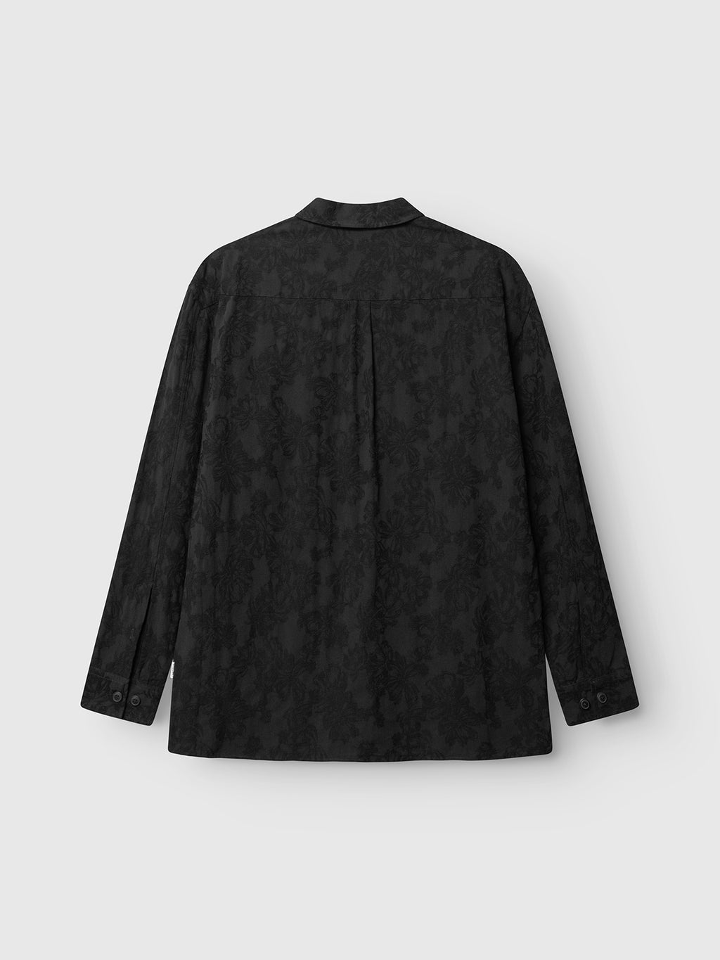 Back view of the GABjens Over Brocade LS Shirt by Gabba, an oversized shirt in black crafted from a cotton-blend fabric with a subtle jacquard floral pattern. Features include buttoned cuffs and a center back pleat. The shirt is displayed against a light gray background.
