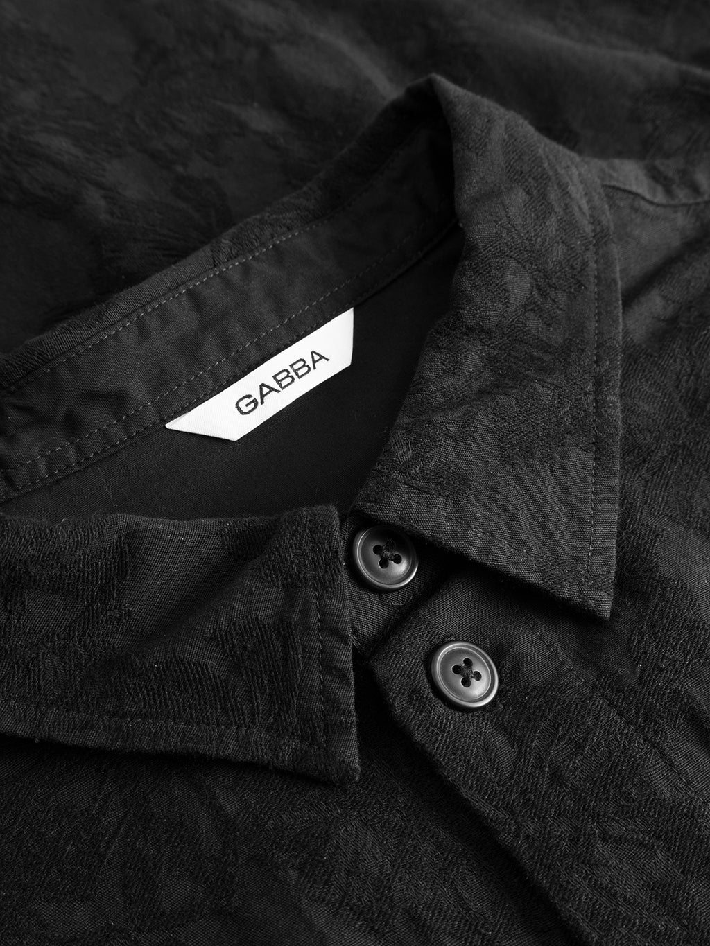 Close-up of the GABjens Over Brocade LS Shirt in black by Gabba, showcasing a black collared design with two buttons and a white tag labeled “Gabba.” The cotton-blend fabric features a subtle jacquard floral textured pattern.