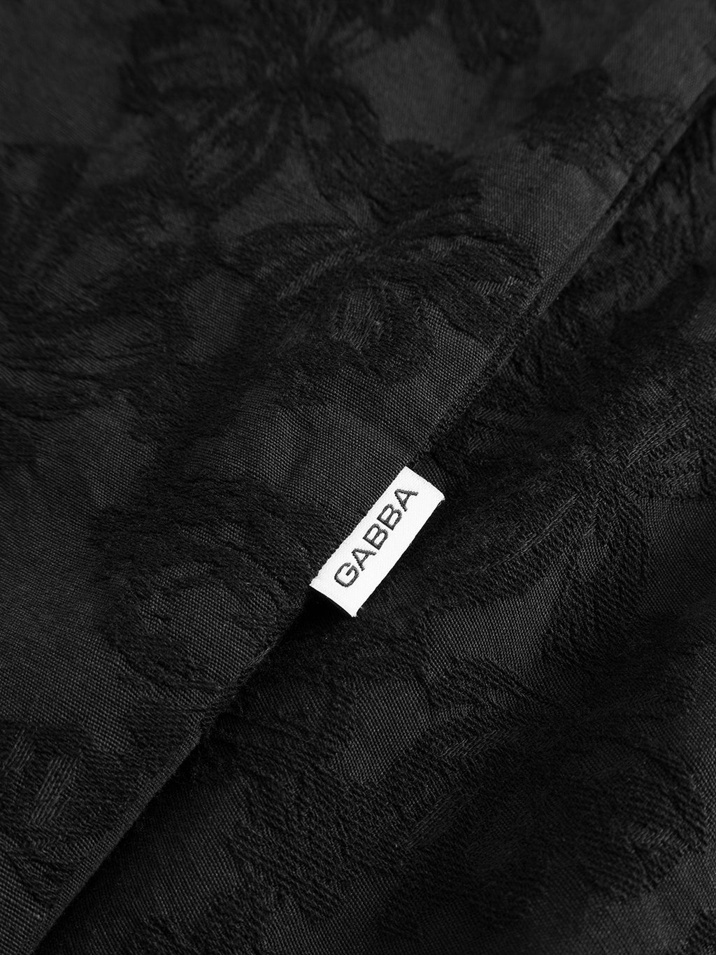 Close-up of the black GABjens Over Brocade LS Shirt by Gabba, showcasing its jacquard floral textured fabric with a subtle pattern and a small white tag labeled “Gabba” sewn into the seam.