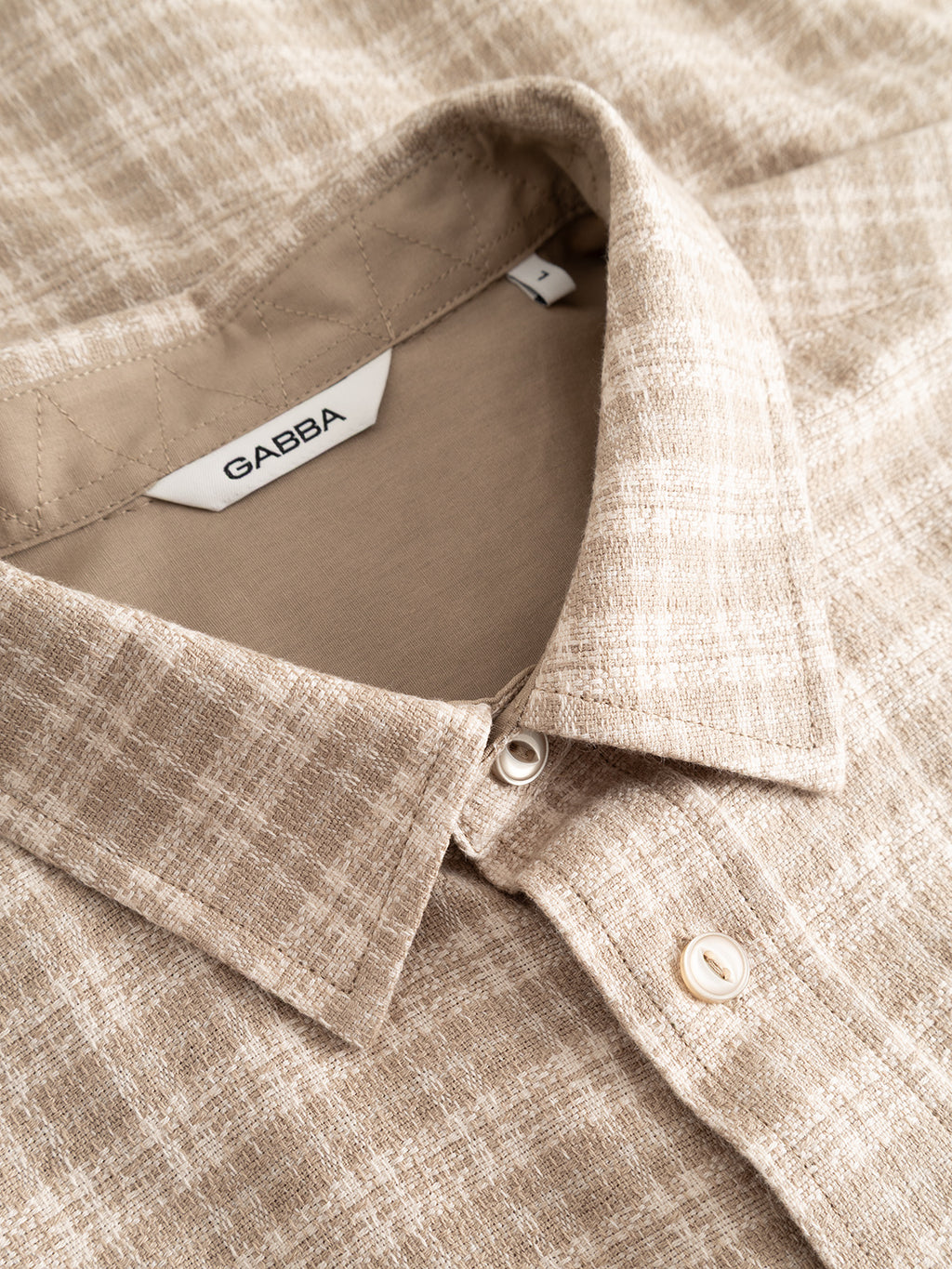 Close-up of the GABsven Reg TX2 LS Shirt - Sand Crocker by Gabba, a beige plaid button-up shirt crafted from yarn-dyed cotton. The regular-fit shirt showcases a visible collar, buttons, and a white tag with the Gabba brand name inside the neckline, featuring a subtle checked texture for timeless style.