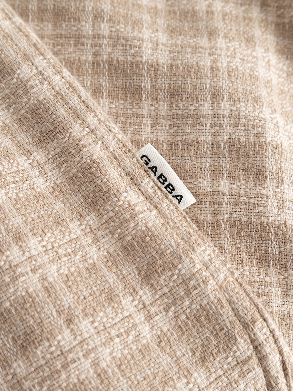A close-up of the sand-colored, yarn-dyed cotton fabric of the GABsven Reg TX2 LS Shirt by Gabba reveals a subtle checked texture and a small white GABBA tag partially tucked into the seam, showcasing the quality and craftsmanship characteristic of this regular-fit shirt.