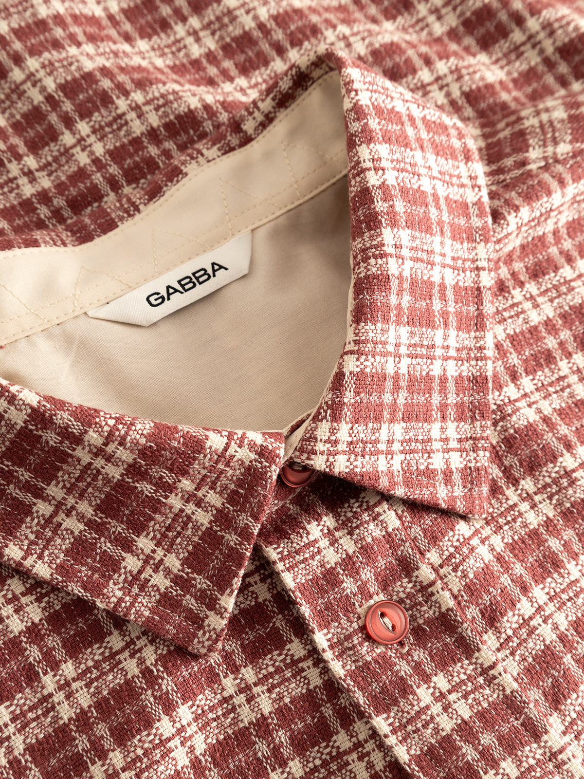 A close-up of the Gabba GABsven Reg TX2 LS Shirt in Brick Red showcases its red and white plaid pattern, regular fit, and two visible buttons. Crafted from yarn-dyed cotton for a striking checked texture, the shirt features a beige inner collar with a fabric label reading “Gabba.”.