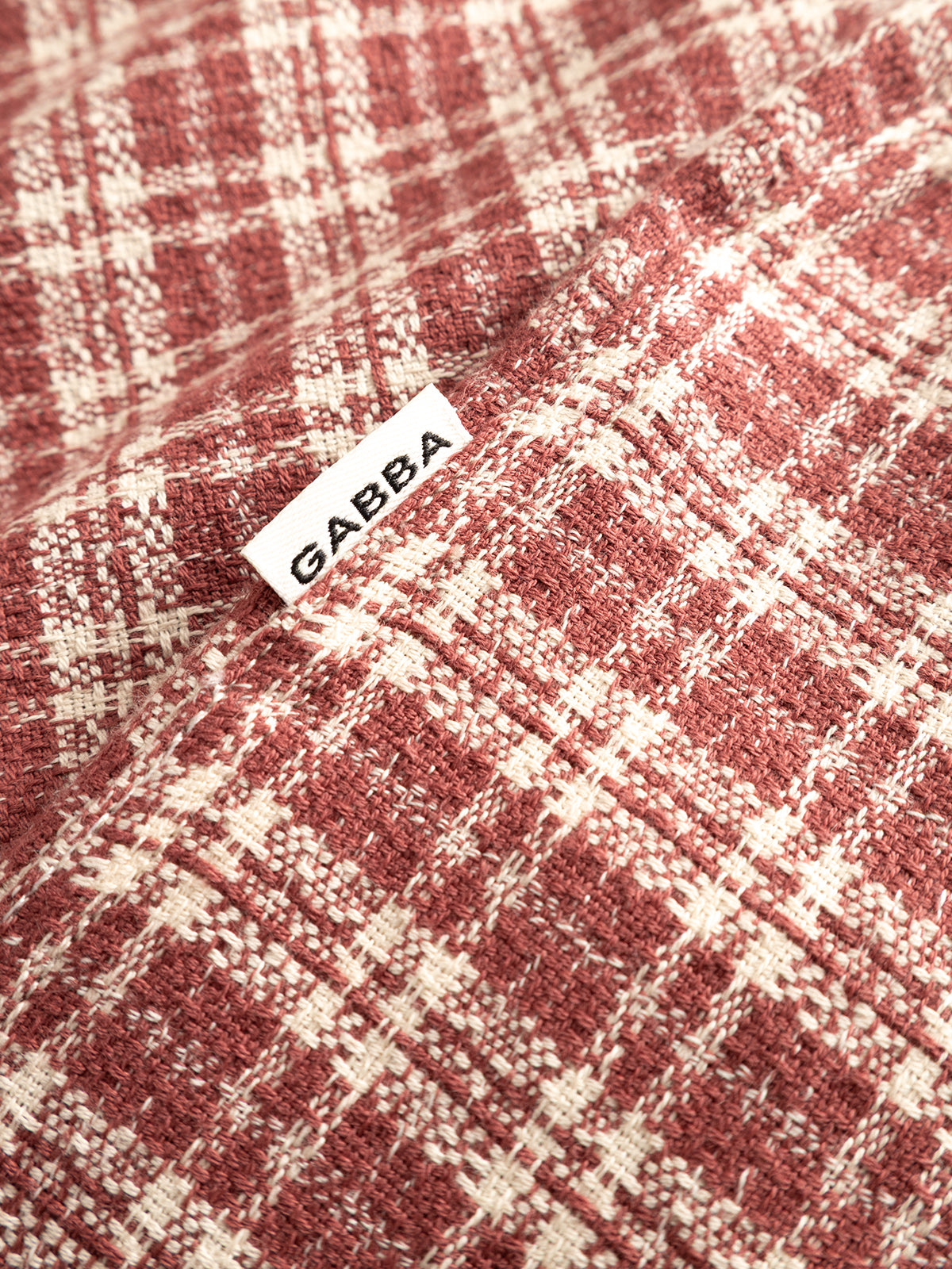 Close-up of the Brick Red GABsven Reg TX2 LS Shirt by Gabba, featuring a red and white checked pattern in yarn-dyed cotton, with a small white GABBA label sewn into the seam.