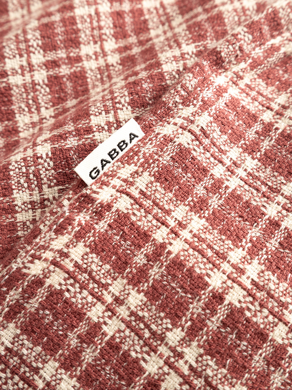 Close-up of the Brick Red GABsven Reg TX2 LS Shirt by Gabba, featuring a red and white checked pattern in yarn-dyed cotton, with a small white GABBA label sewn into the seam.