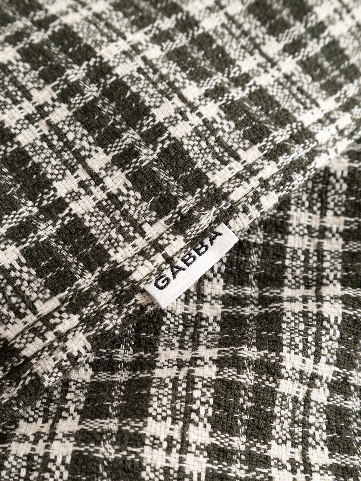 Close-up of overlapping black and white plaid patterns crafted from yarn-dyed cotton fabric, showcasing a subtle checked texture. A visible white tag displays the brand name Gabba in bold black letters. Shown: GABsven Reg TX2 LS Shirt - Forest Night by Gabba.