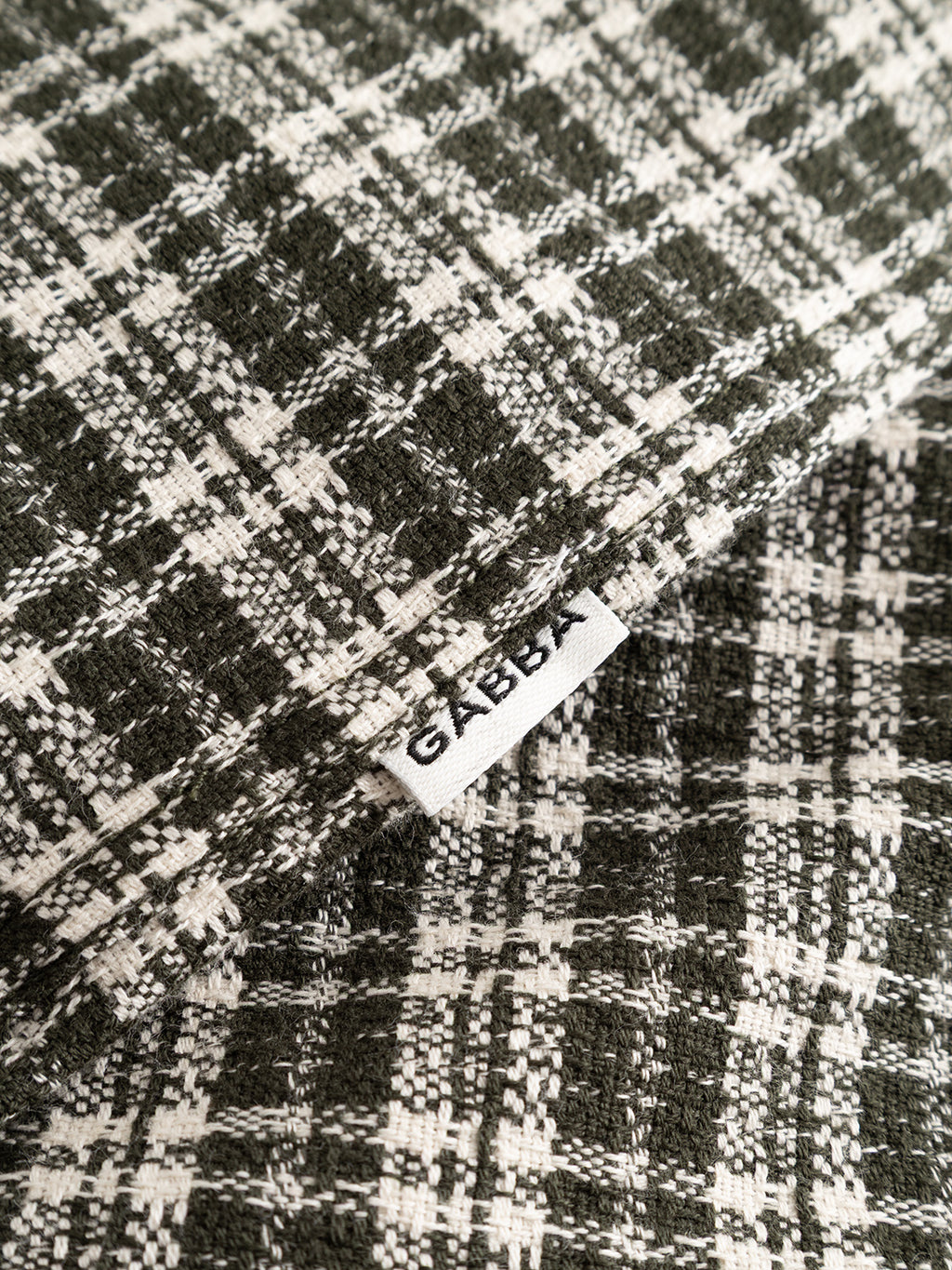 Close-up of overlapping black and white plaid patterns crafted from yarn-dyed cotton fabric, showcasing a subtle checked texture. A visible white tag displays the brand name Gabba in bold black letters. Shown: GABsven Reg TX2 LS Shirt - Forest Night by Gabba.