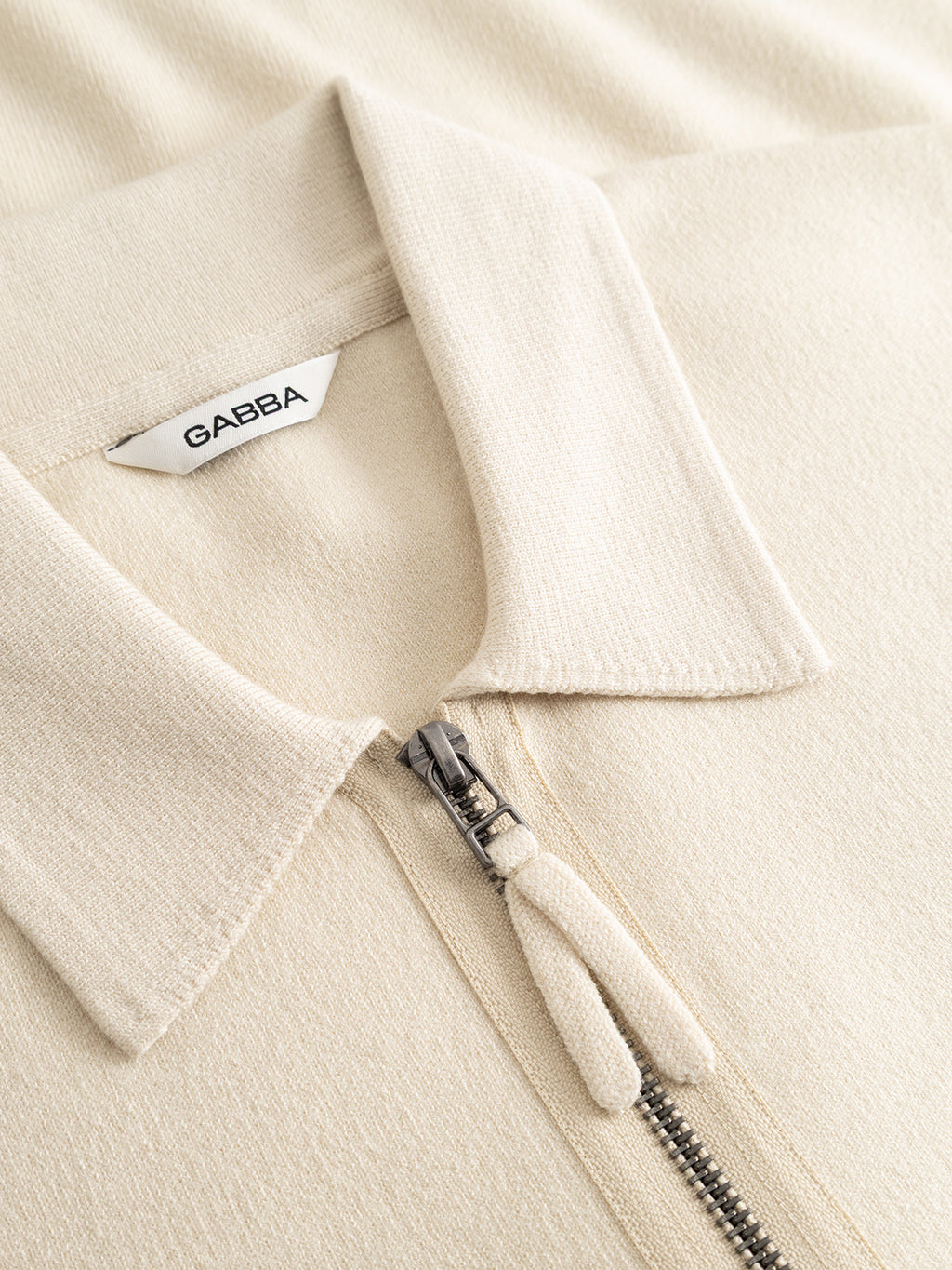Close-up of the GABadriel Zip Lume LS Knit in Birch by Gabba—a beige collared shirt crafted from a soft, textured viscose blend knit. It features a metal full zip closure and a white tag with the Gabba logo.