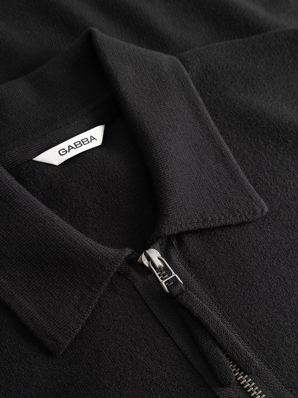 Close-up of the GABadriel Zip Lume LS Knit - Meteorite by Gabba, a black collared shirt with a full zip closure and a white “GABBA” label inside the collar, crafted from a soft viscose blend for enhanced comfort.