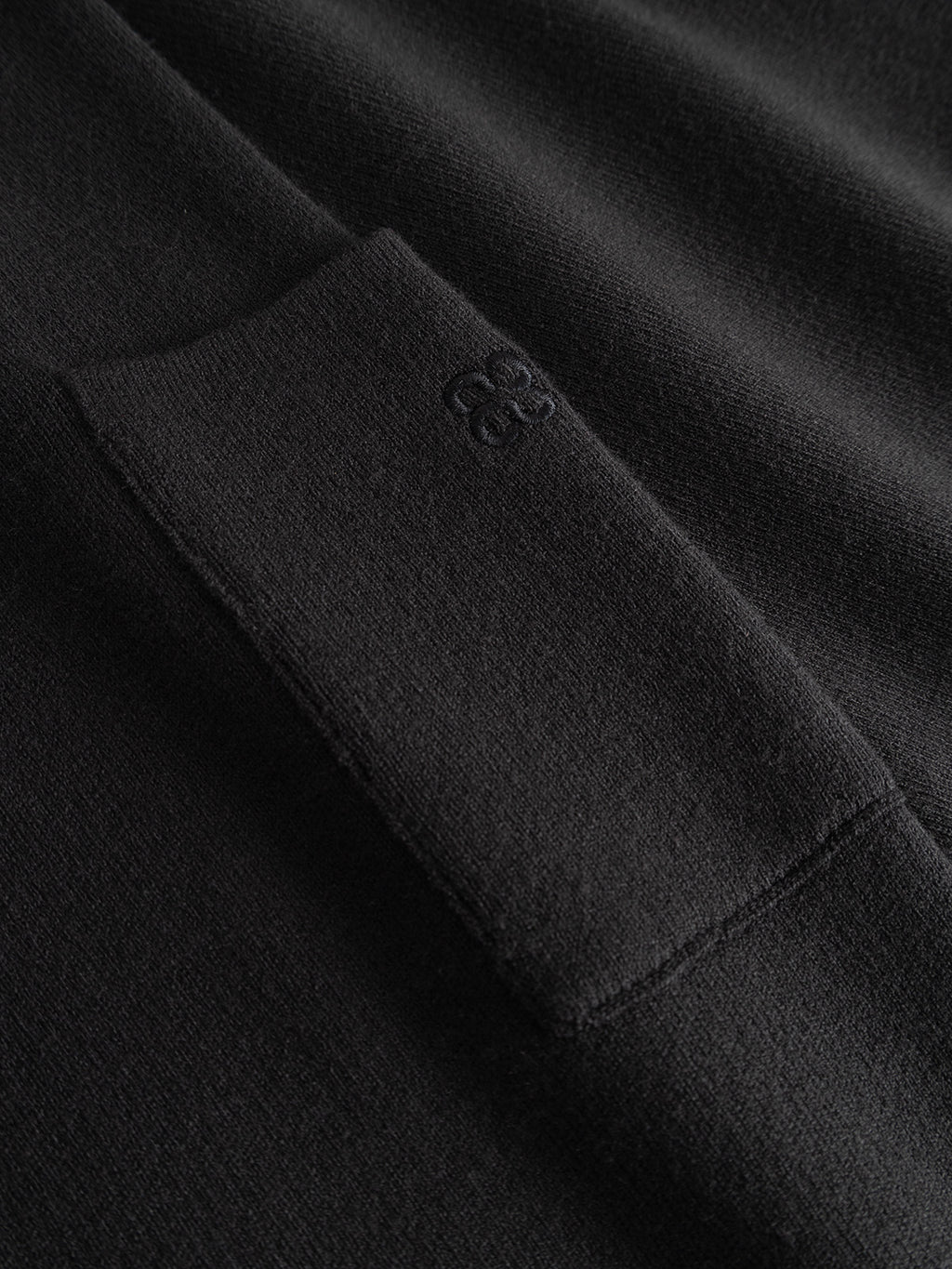 Close-up of the GABadriel Zip Lume LS Knit - Meteorite by Gabba, featuring black regular fit knit fabric with a small rectangular pocket and subtle embroidered logo, highlighting the texture and stitching details.