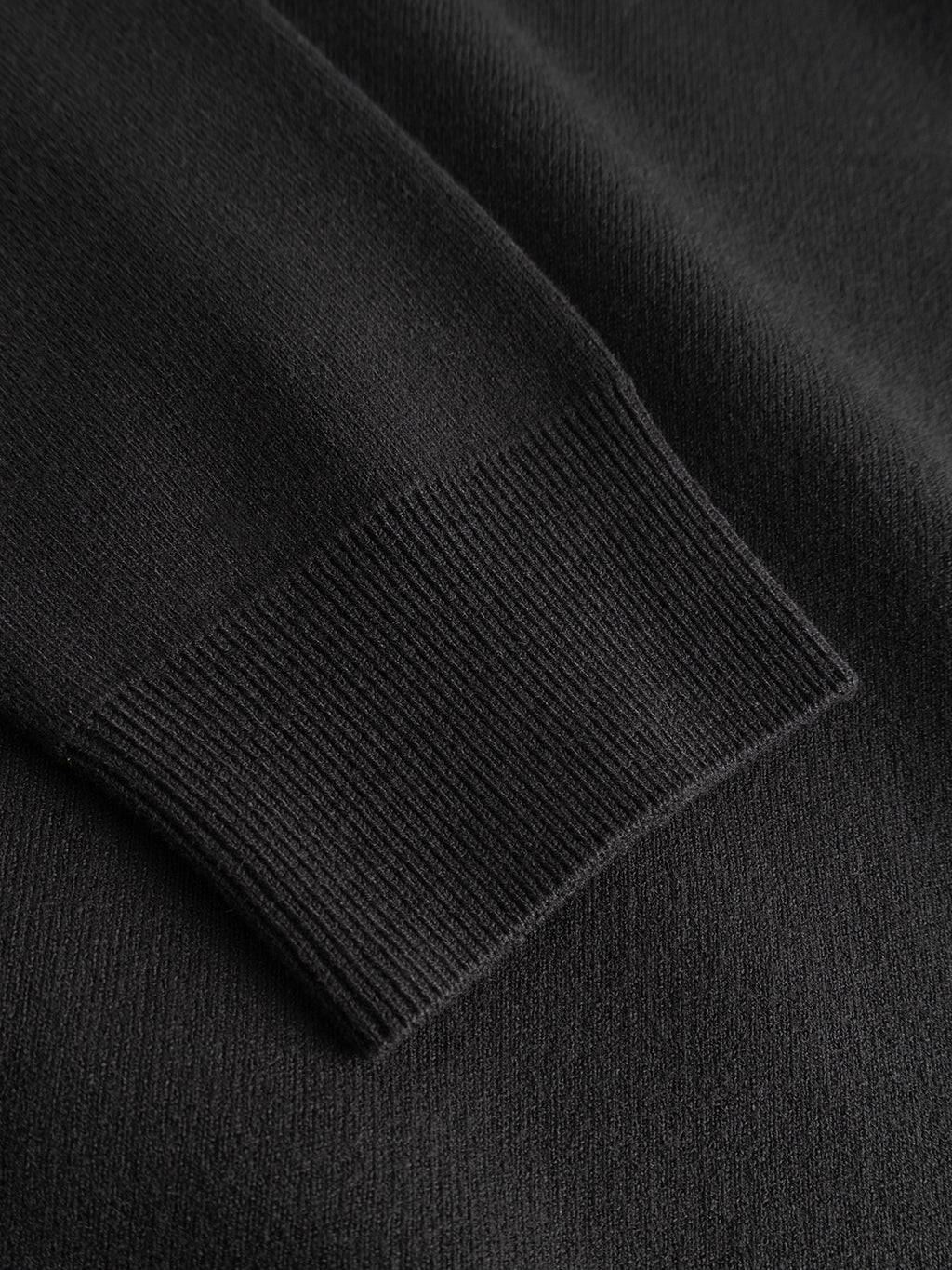 Close-up of the GABadriel Zip Lume LS Knit - Meteorite by Gabba, featuring a black regular-fit sleeve with a ribbed cuff, laid flat on a matching black surface.