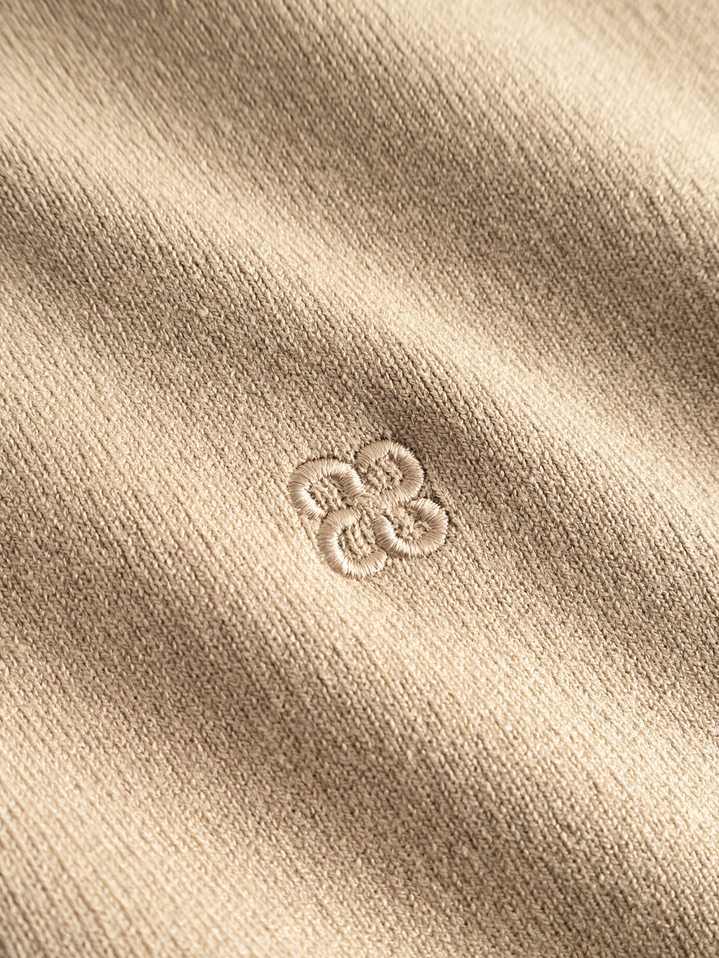 Close-up of the Gabba GABadriel Half Zip Lume LS Knit in Sand Crocker, showcasing its beige viscose blend fabric. A small, embroidered abstract floral or geometric logo design appears at the center in matching thread, highlighting the texture of this regular fit knit.