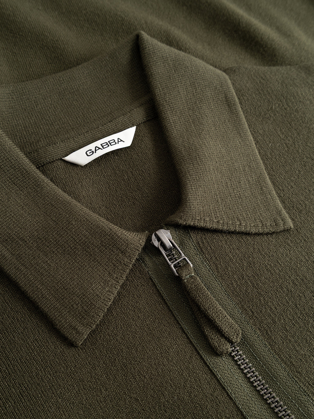 A close-up of the GABadriel Half Zip Lume LS Knit in Forest Night by Gabba shows its olive green, collared half-zip sweater design with a white Gabba tag under the collar; the soft, textured fabric is crafted from a viscose blend knit.