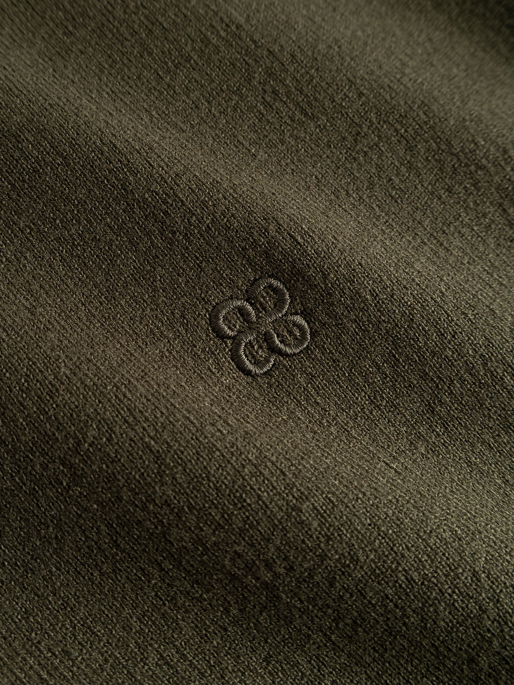 Close-up of the “GABadriel Half Zip Lume LS Knit - Forest Night” by Gabba, showcasing olive green fabric with a subtle textured weave and a small, tonal embroidered logo centered on the chest—resembling stylized interlocking loops or a four-leaf clover. Crafted from a soft viscose blend knit, this regular fit half-zip sweater blends refined detailing with everyday comfort.