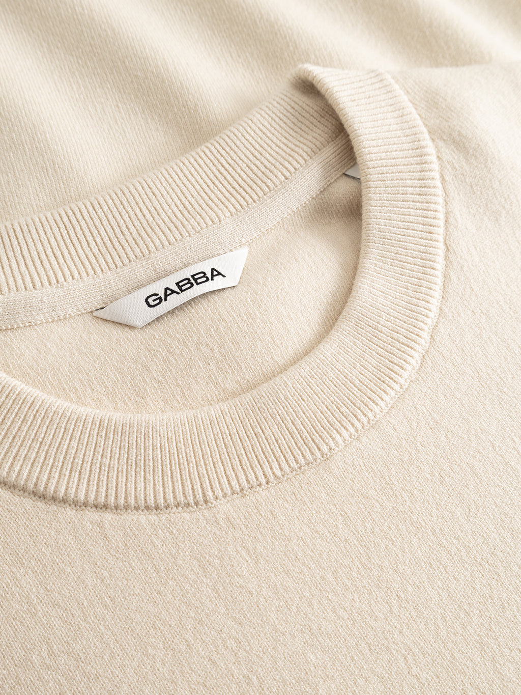 Close-up of the GABadriel O-Neck Lume LS Knit - Birch by Gabba, a beige knit sweater featuring a ribbed crew neck collar and a white label inside the neckline that reads Gabba. Crafted from a soft viscose blend, this regular fit knit offers both comfort and timeless style.