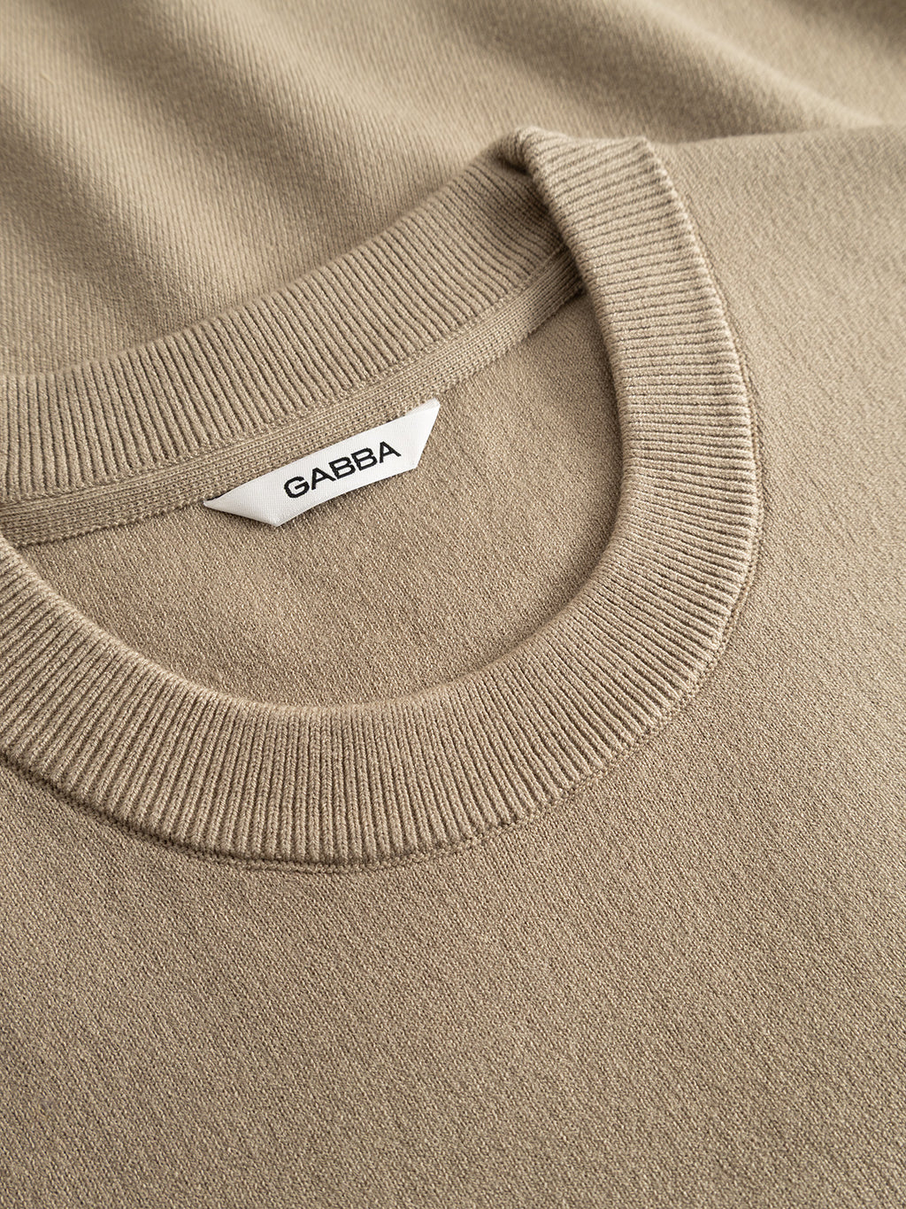 Close-up view of the ribbed crew neck of the GABadriel O-Neck Lume LS Knit - Sand Crocker, a beige viscose blend sweater, featuring a white garment tag with the Gabba brand name printed in black.