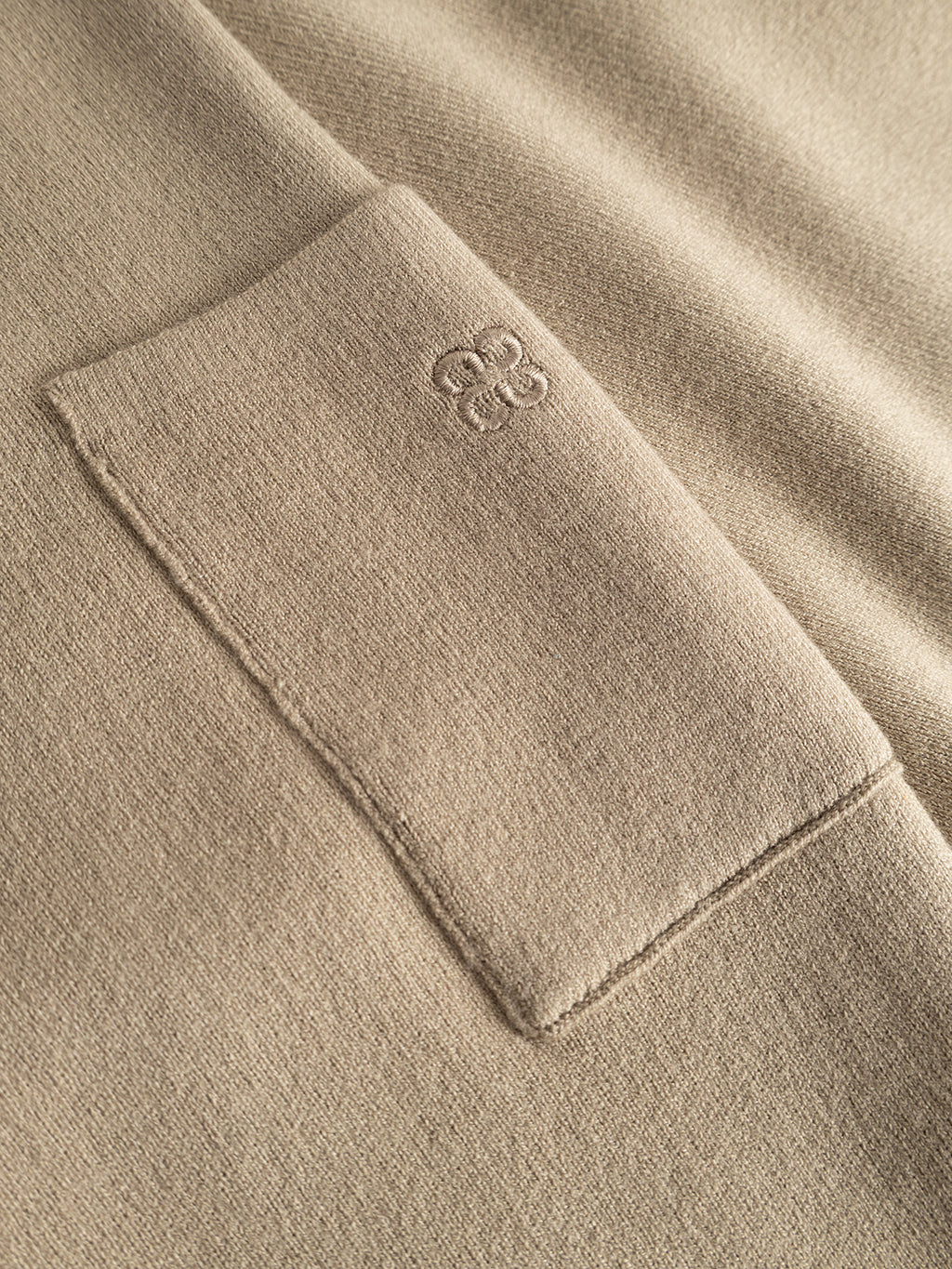 A close-up of the Gabba GABadriel O-Neck Lume LS Knit in Sand Crocker showcases its beige viscose blend fabric and a square chest pocket. The pocket is adorned with an embroidered Gabba logo of four interlocking circles near the top edge. The regular fit, crew neck sweater design is highlighted by lighting that emphasizes the garment’s subtle texture and shadows.