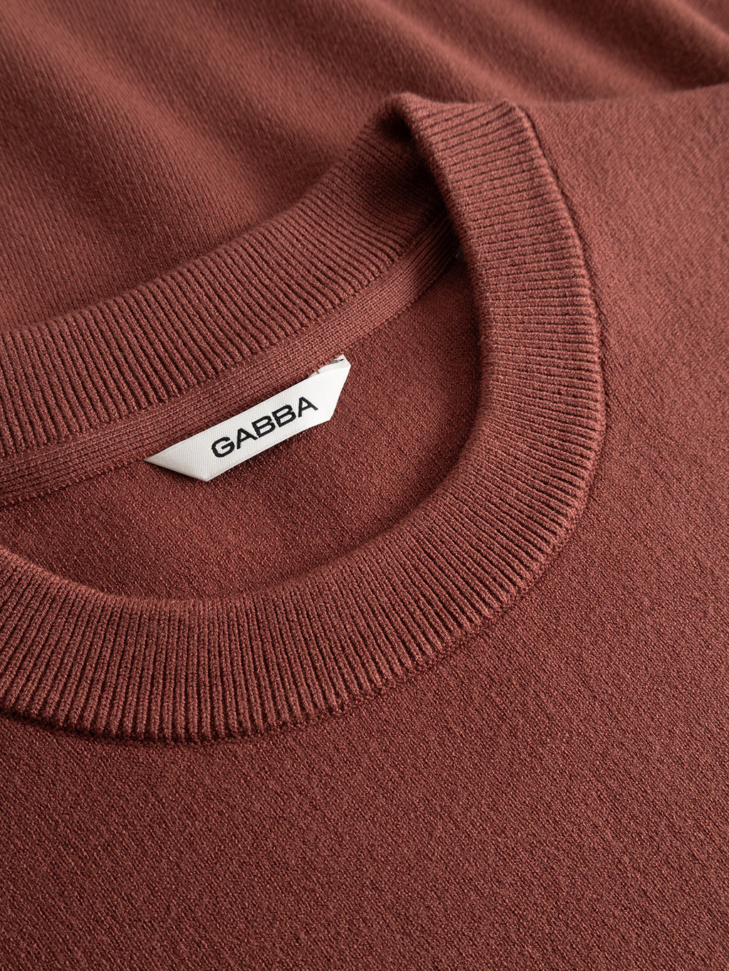 A close-up of the GABadriel O-Neck Lume LS Knit in Brick Red by Gabba, a regular fit sweater crafted from a soft viscose blend. It features a ribbed crew neckline and has a white “GABBA” label sewn inside the collar.