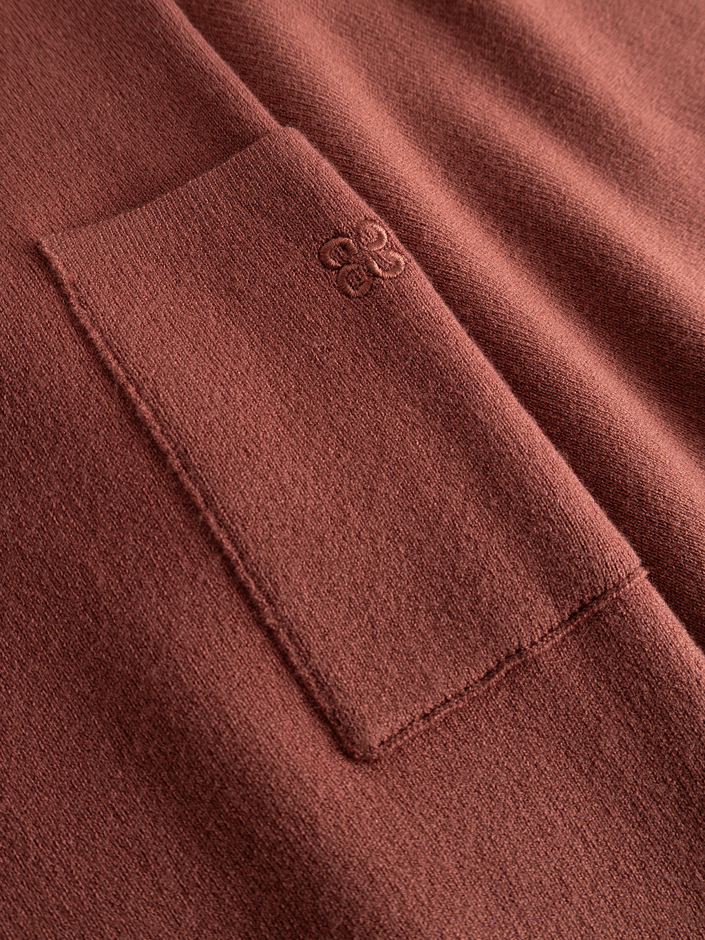 Close-up of the GABadriel O-Neck Lume LS Knit in Brick Red by Gabba, featuring a textured viscose-blend fabric with a rectangular pocket and an embossed floral logo atop the pocket.