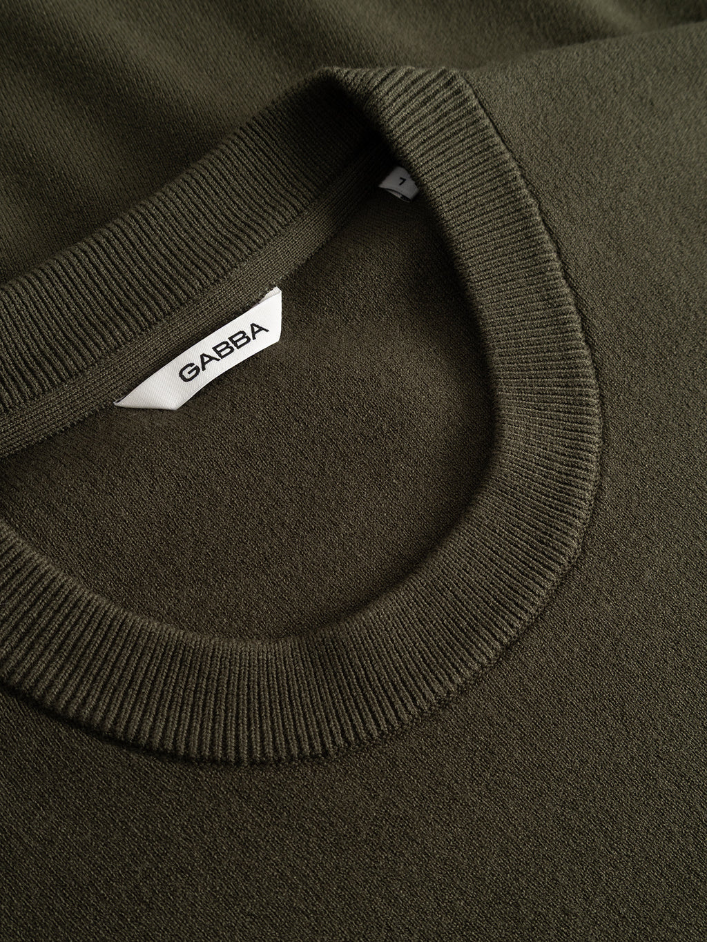 Close-up of the GABadriel O-Neck Lume LS Knit in Forest Night, a regular-fit crew neck sweater by Gabba crafted from a mid-weight viscose blend. The photo highlights the texture and stitching of the knit fabric, with a visible white label inside the collar displaying the brand name Gabba.