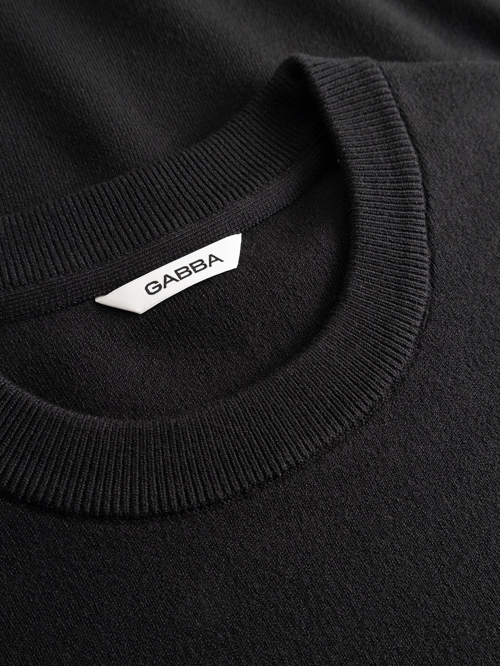 Close-up of the GABadriel O-Neck Lume LS Knit - Meteorite, a black crew neck sweater from Gabba. This regular-fit sweater is crafted from a viscose blend knit and features a ribbed neckline. A white tag inside the collar displays the brand name “Gabba” in black uppercase letters. The texture of the fabric is clearly visible.