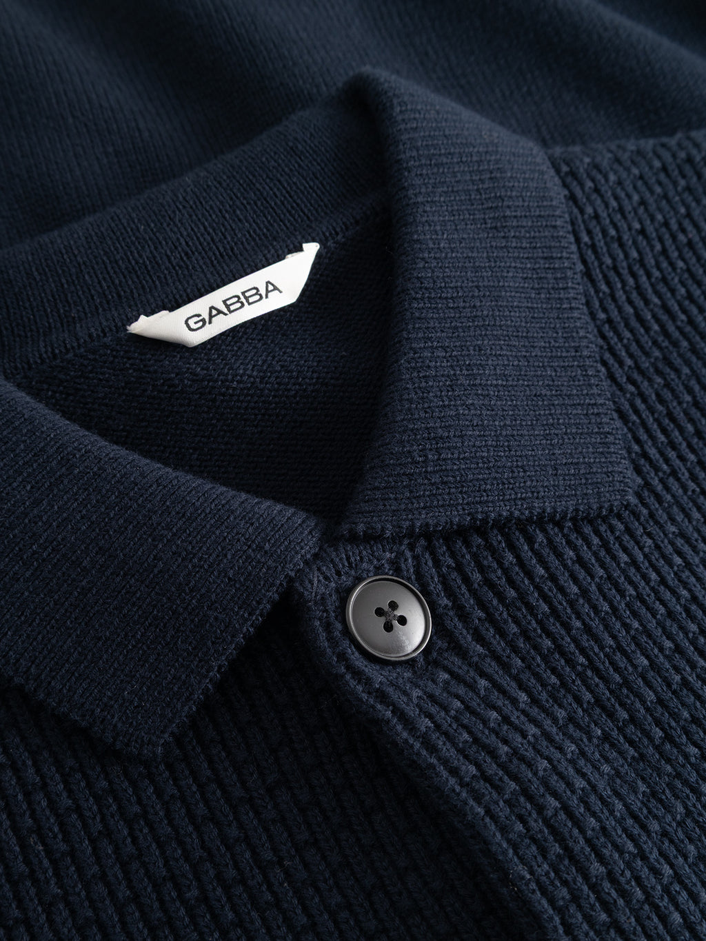 Close-up of the GABbenoit Cardi Fort LS Knit - Night Sky by Gabba, featuring a dark navy structured knit with a collar, a single black button, and a white GABBA tag, highlighting its regular fit design.