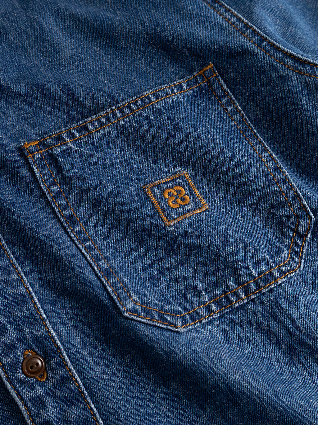 Close-up of the GABsven Reg Wish LS Shirt by Gabba in mid blue denim, featuring a cotton pocket with orange stitching and a small embroidered square logo at the center. A brown button is partially visible on the left, highlighting the versatility and detailed craftsmanship of this essential piece.