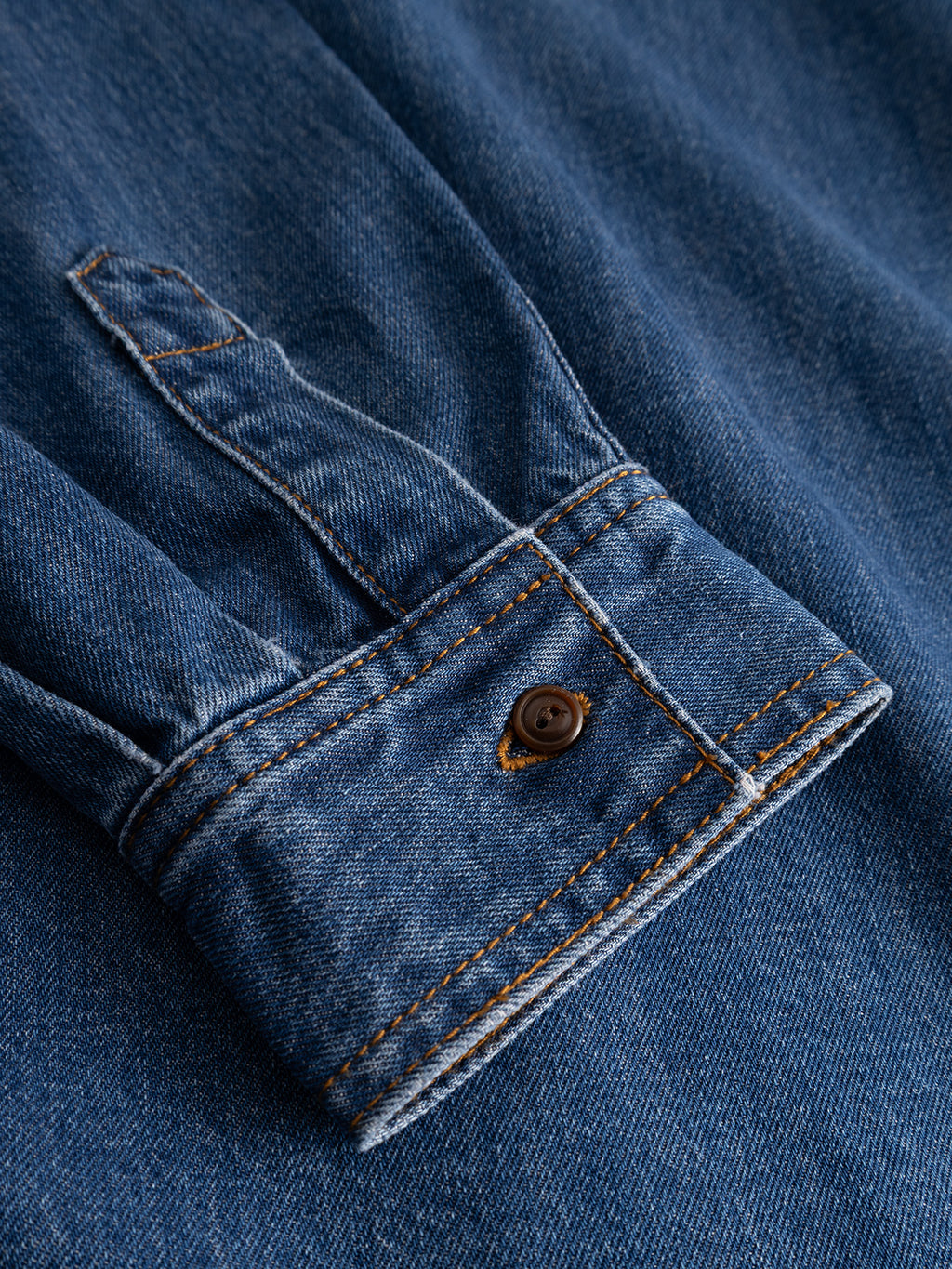 Close-up of the GABsven Reg Wish LS Shirt by Gabba in mid blue denim, featuring a textured cotton fabric with orange stitching and a brown button at the cuff. The detailed seams and slightly faded look make this shirt a versatile wardrobe essential for any occasion.