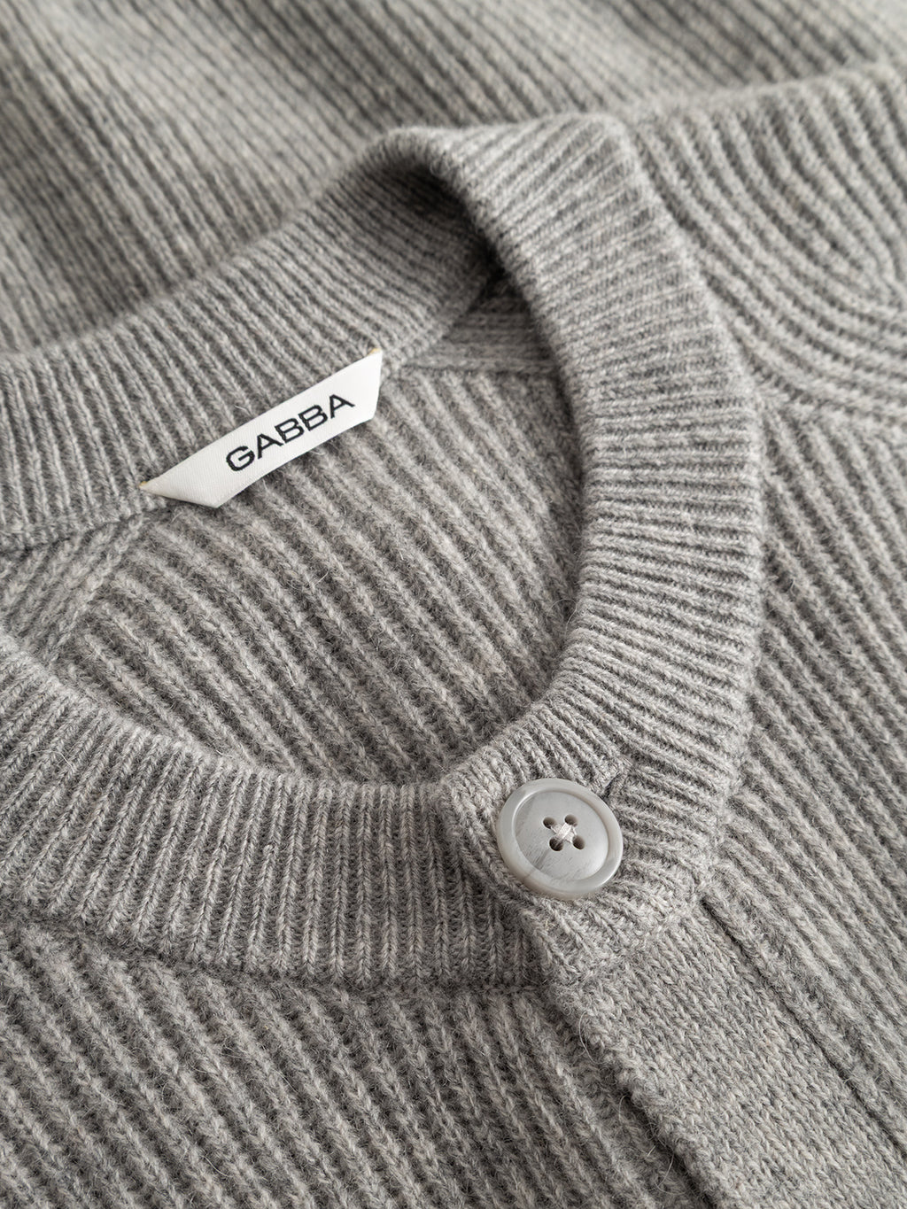 A close-up of the Gabba GABremy Cardi Flash LS Knit in Grey Mel showcases its rib-knit wool texture, round neckline, single button closure, and white Gabba label. The relaxed fit enhances its casual yet refined look.